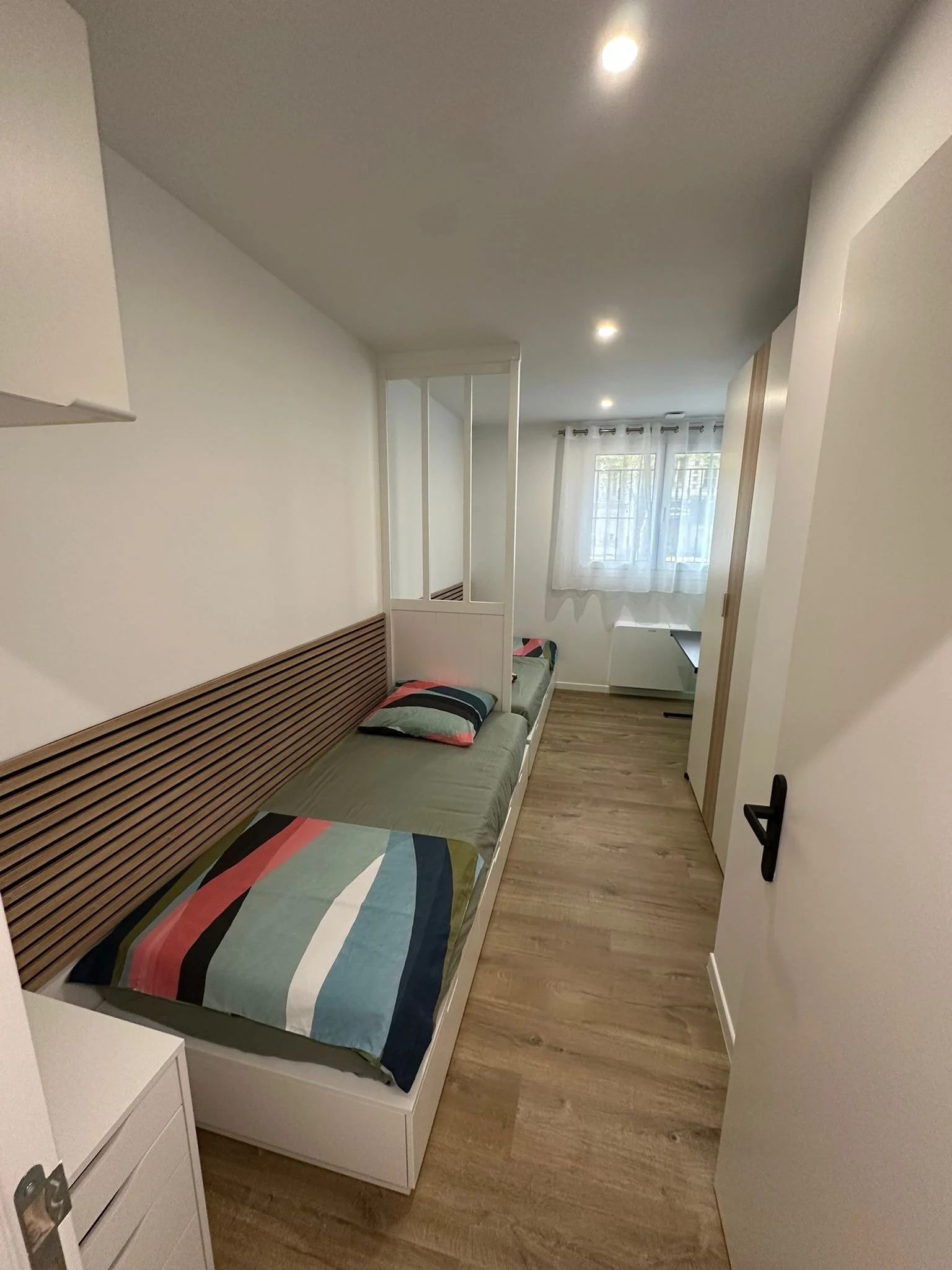  chambre avec un lit simple recouvert d'une couverture à rayures colorées, un coussin, une fenêtre avec rideaux blancs, un sol en bois, et un mur blanc, tasseaux séparateur