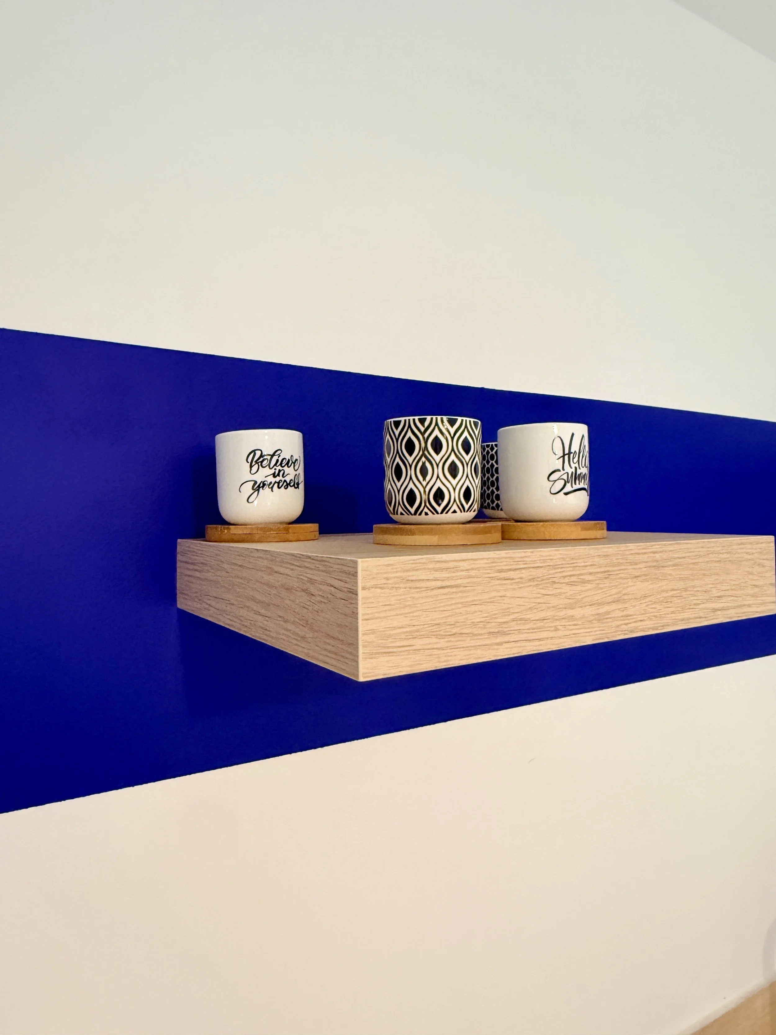 Trois mugs blancs avec des phrases en anglais, placés sur des dessous de verre en bois, posés sur une étagère en bois au mur avec une peinture bicolore bleue et blanche.