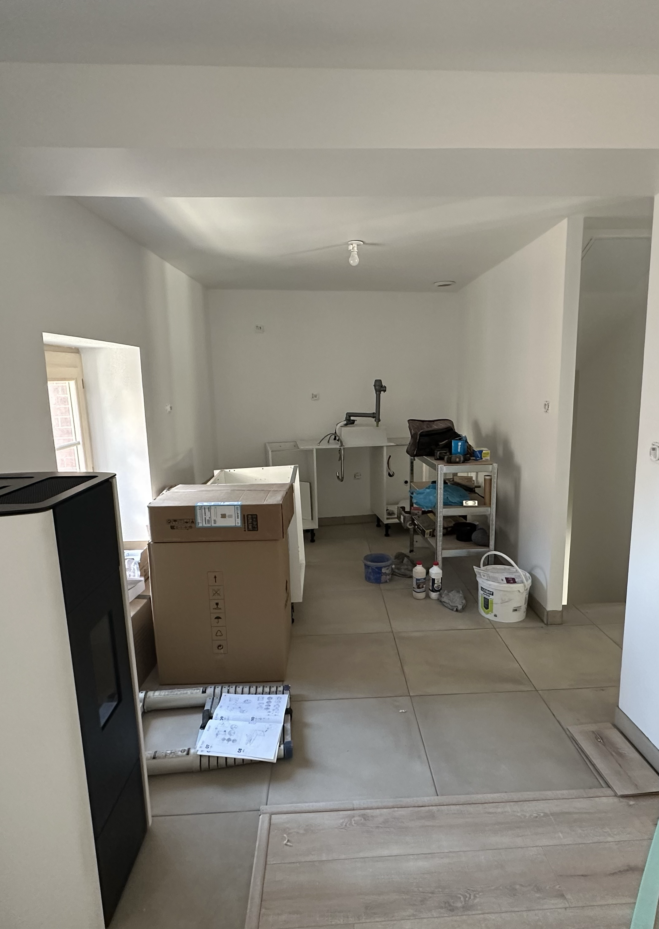 Une pièce en cours d'aménagement avec des meubles, des boîtes, des outils et des matériaux de rénovation. La pièce est lumineuse avec des murs blancs et un sol en carrelage beige.