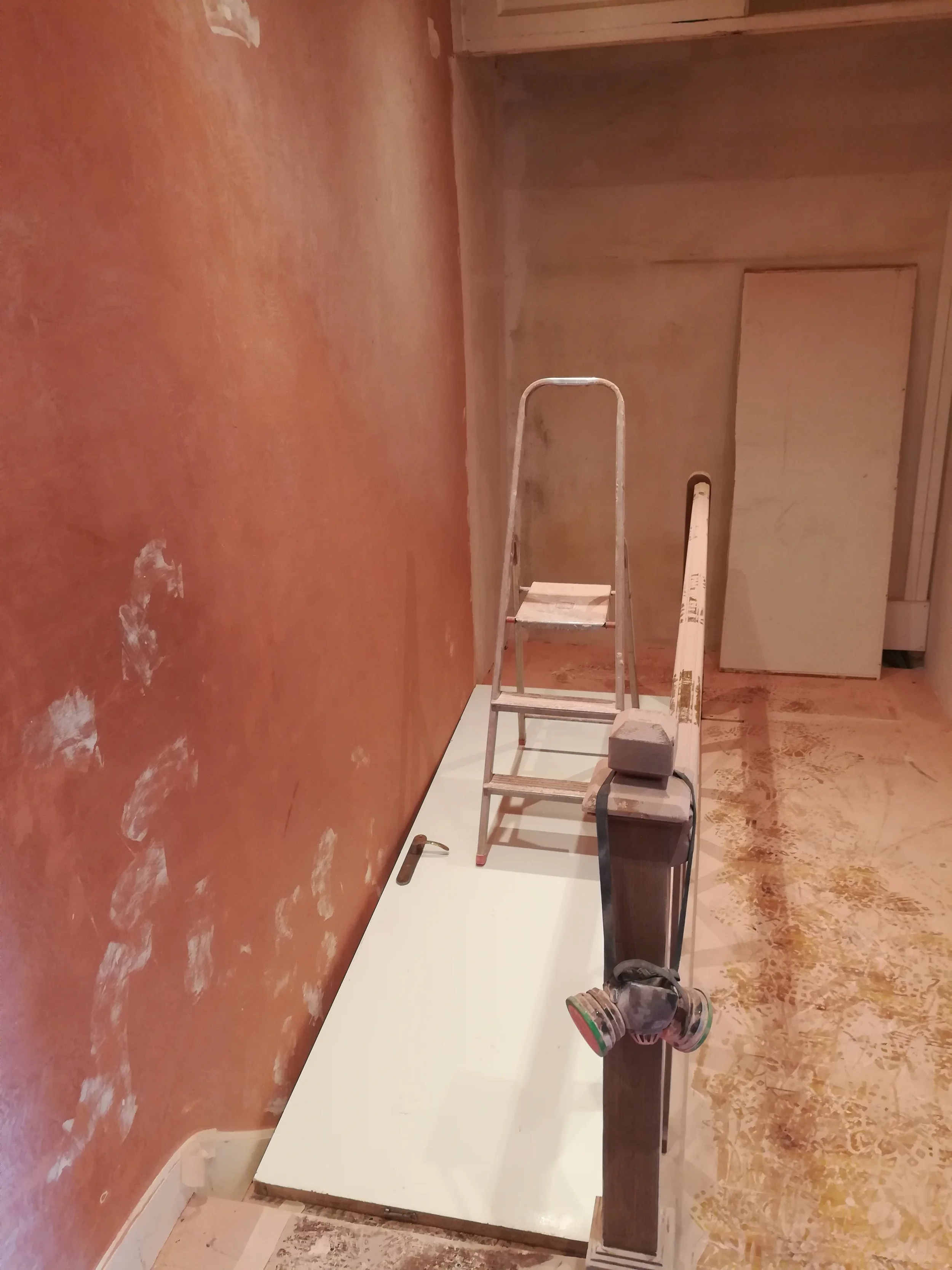 Une pièce en travaux avec un mur peint en rouge, un escabeau en métal, un rouleau de peinture, une planche en bois posée sur un meuble, et un panneau ou placard beige en arrière-plan.