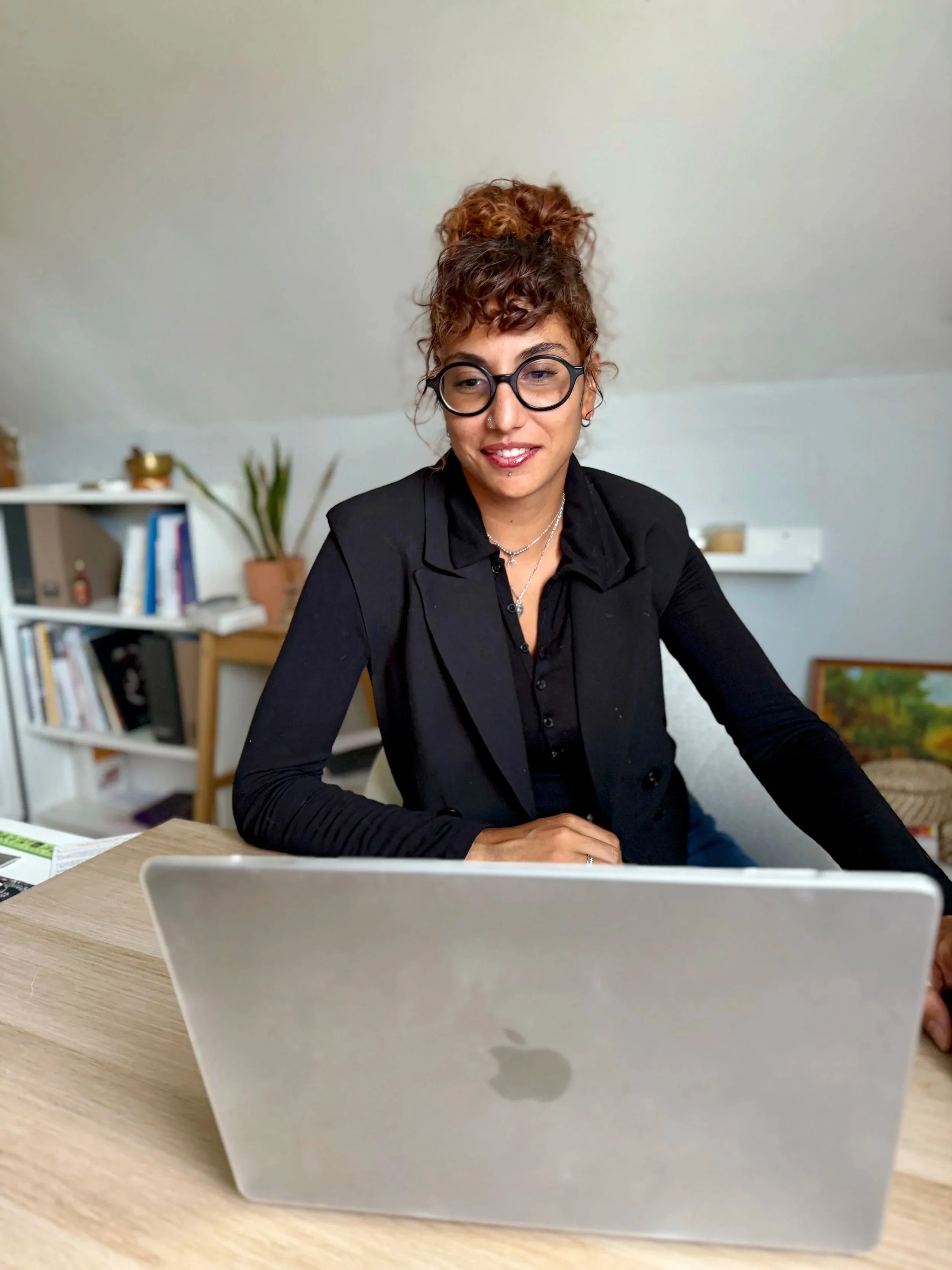Une femme avec des cheveux roux en chignon, portant des lunettes rondes et un collier, assise à une table d'intérieur, avec un ordinateur en premier plan et une étagère en arrière-plan.