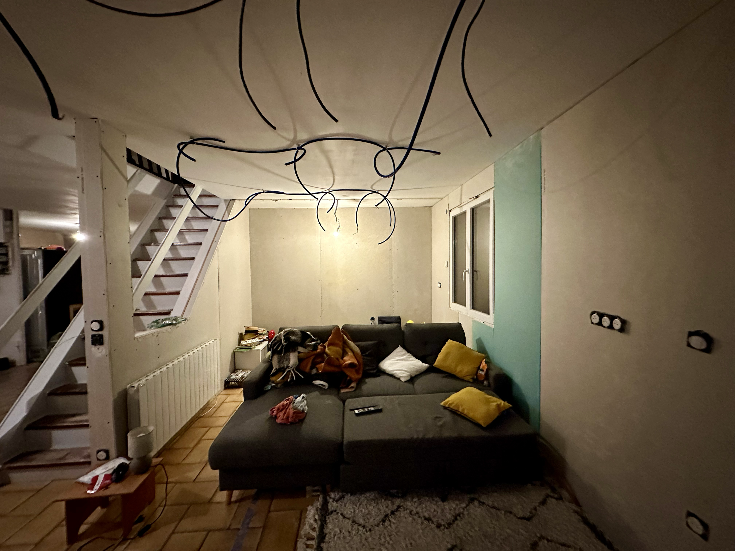 Salon de séjour avec un canapé noir, des coussins jaunes, un tapis, un meuble TV avec des livres et des objets, et un escalier en bois menant à une mezzanine, dans une pièce partiellement en cours de rénovation avec des câbles électriques suspendus a