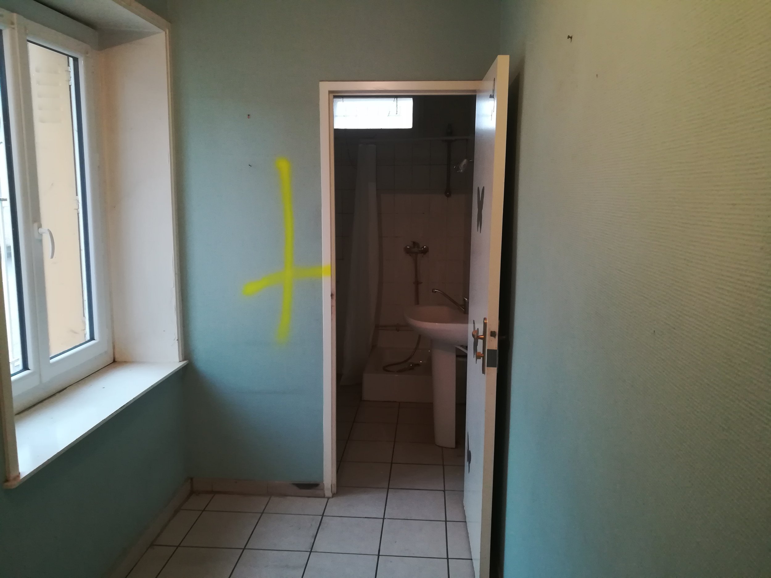 Une pièce intérieure avec un mur vert clair, une fenêtre à gauche, une porte blanche ouverte menant à une salle de bain ou buanderie, avec un lavabo, une plomberie visible, et un sol carrelé. Il y a une marque en jaune de peinture ou sur le mur.