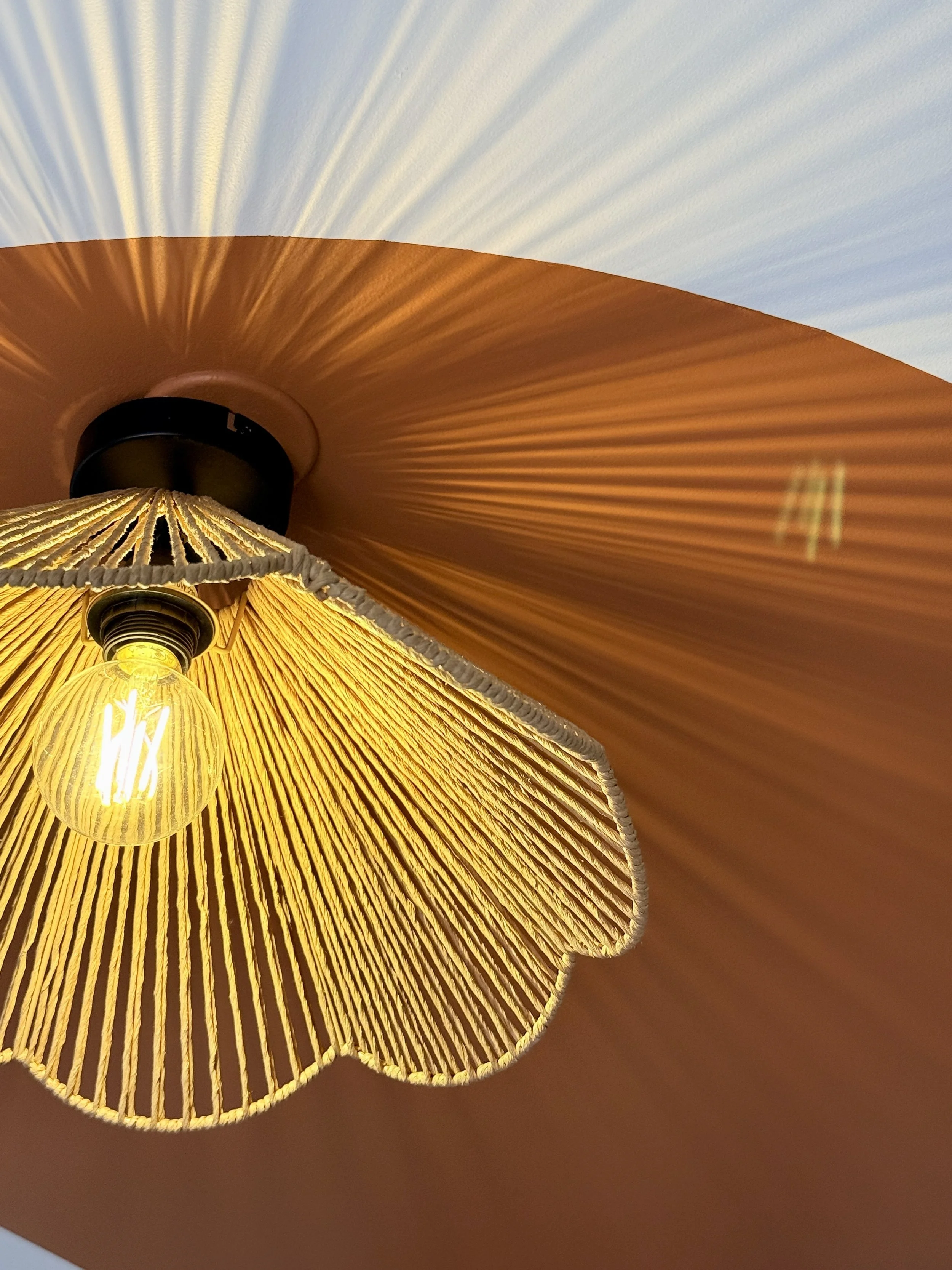 Chandelle suspendue illuminant avec un abat-jour en papier teinté orange et un autre en tissu blanc, avec un fond de ciel bleu clair.