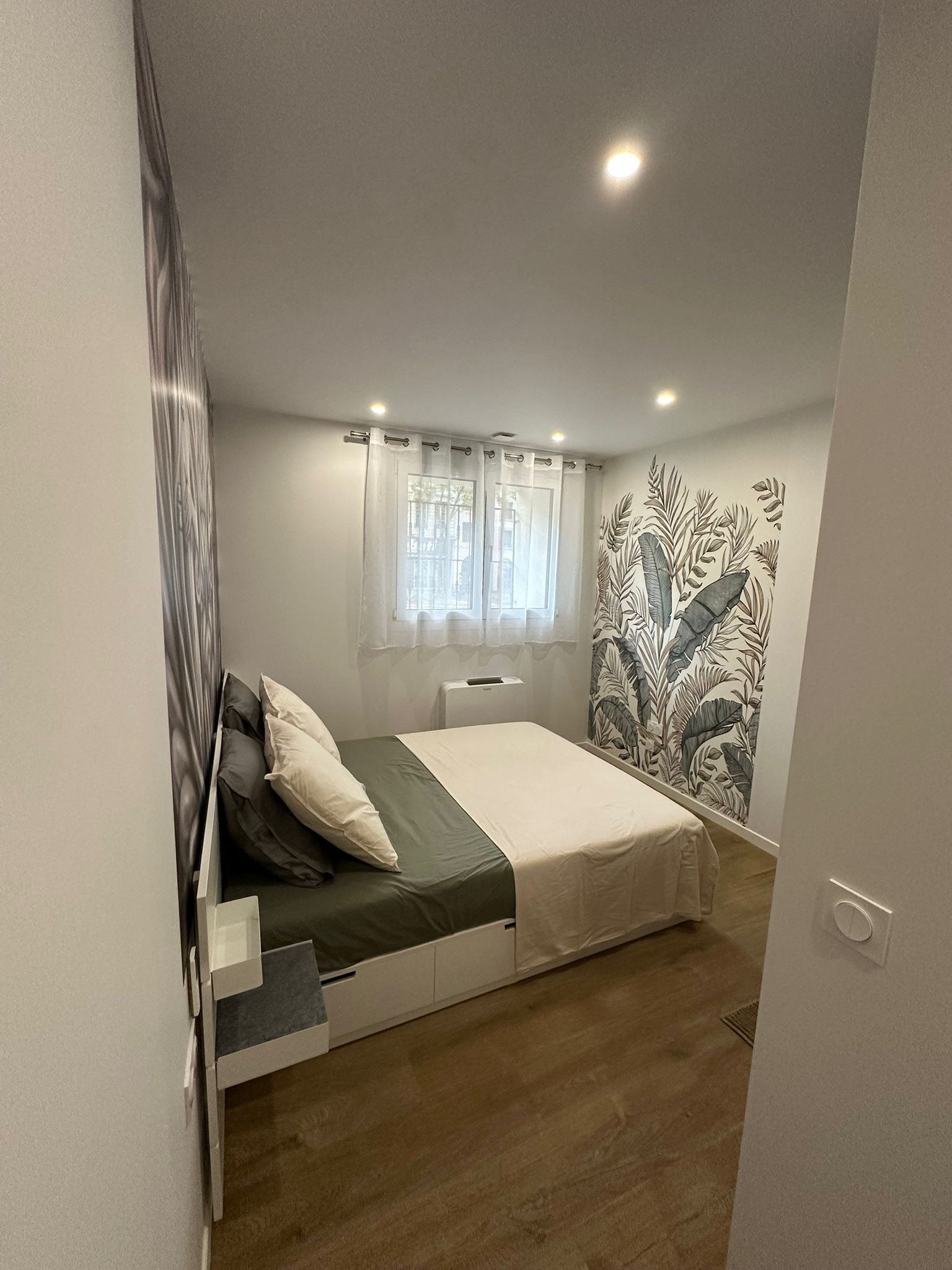 Chambre moderne avec un lit double, des oreillers gris et blancs, une fenêtre avec rideaux blancs, un mur décoratif avec un motif végétal, et un plafond avec des spots lumineux.