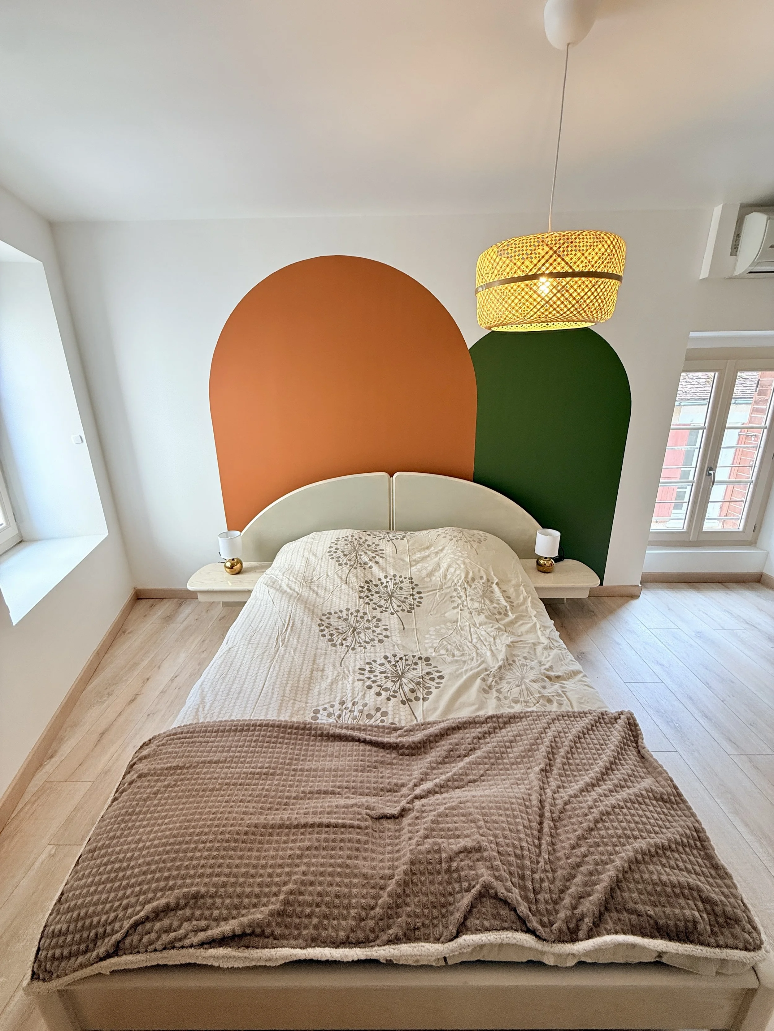 Chambre avec lit en bois clair, une tête de lit blanche, deux petits lampes de chevet dorées, un mur avec une décoration en forme de deux arches colorées, une grande lampe suspendue avec un abat-jour jaune à motif.