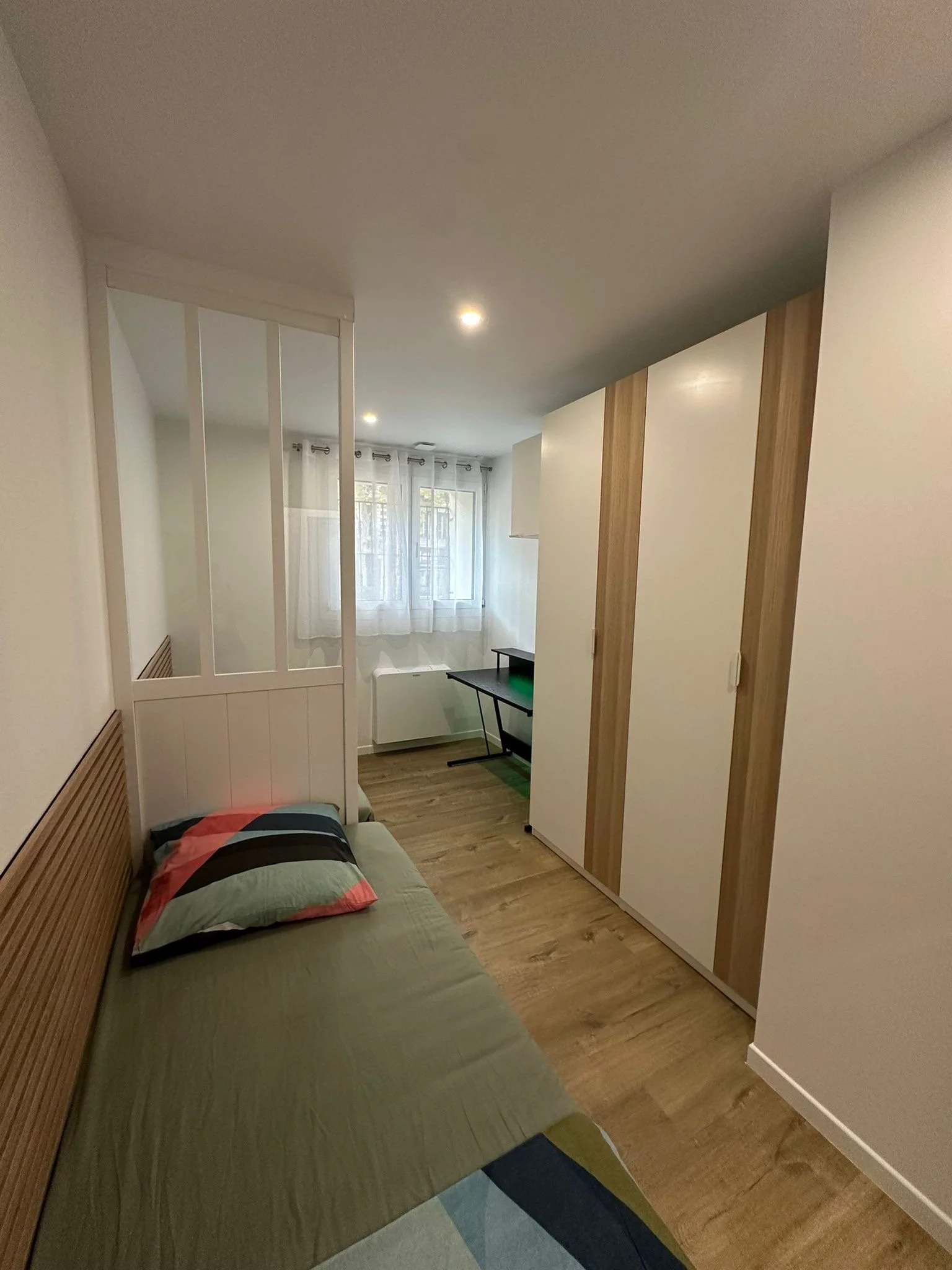 Chambre avec un lit simple, un coussin à rayures, un placard avec portes blanches et en bois, une fenêtre avec rideaux lumineux, et du sol en bois lamellé.