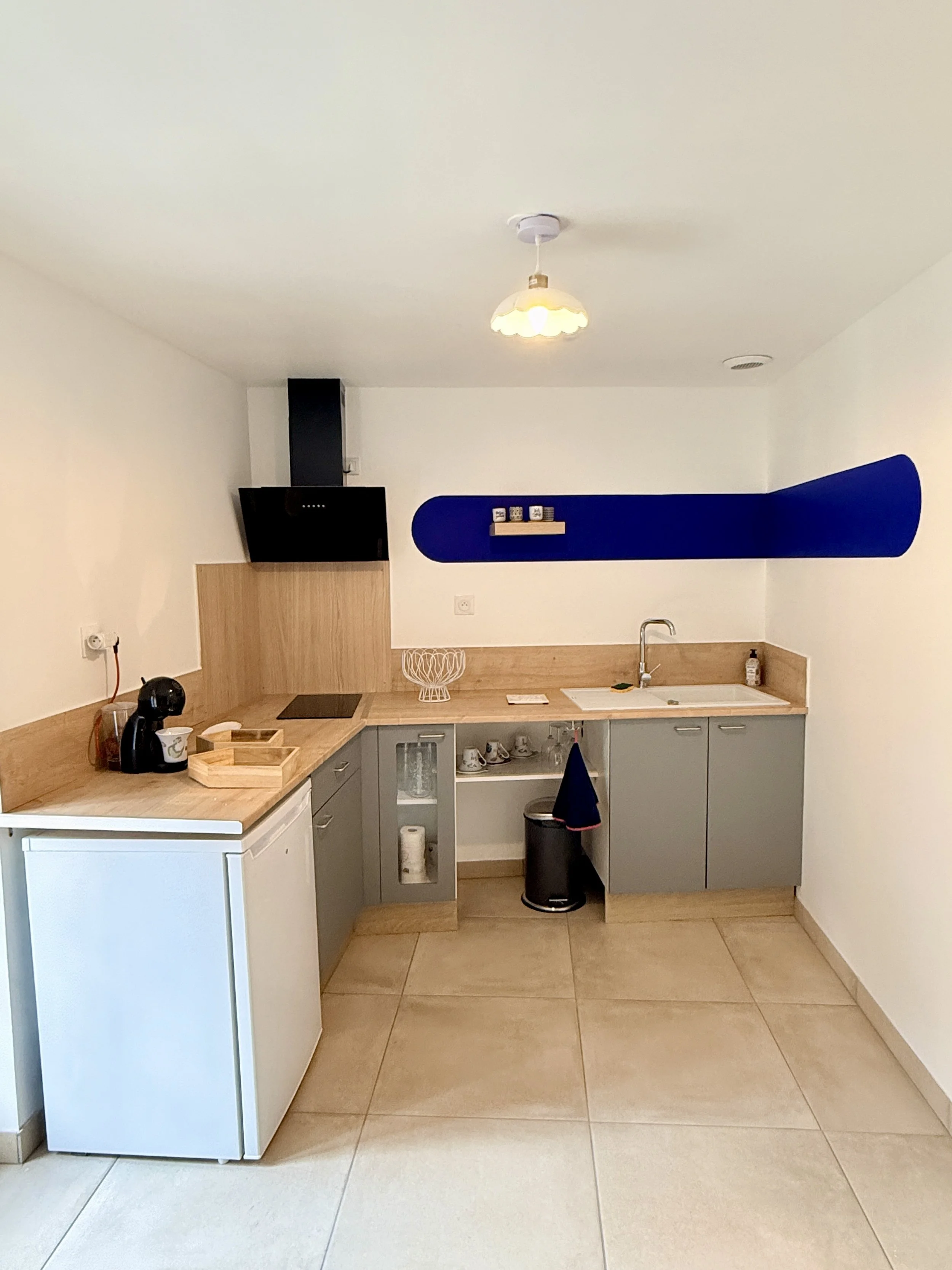 Cuisine moderne avec des armoires grises, un évier, un petit réfrigérateur blanc, un lave-vaisselle, une étagère avec des tasses, une planche à découper et un porte-rouleau. Mur blanc avec une pièce de décoration bleue en forme de chemin. Un plafonni