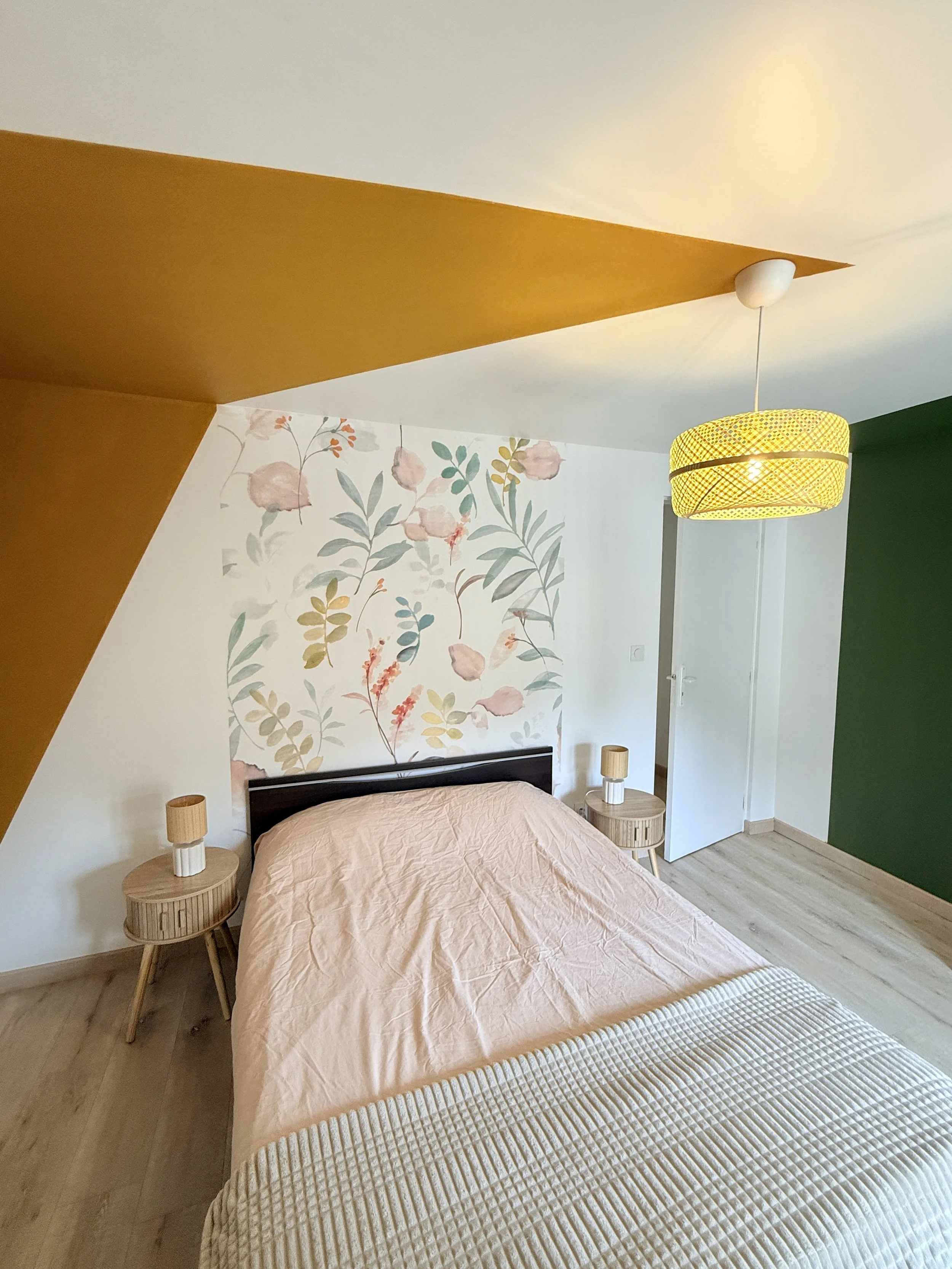 Chambre avec lit en bois, deux tables de chevet en bois avec lampes, mur avec papier peint floral, suspension en rotin jaune, mur vert à droite.