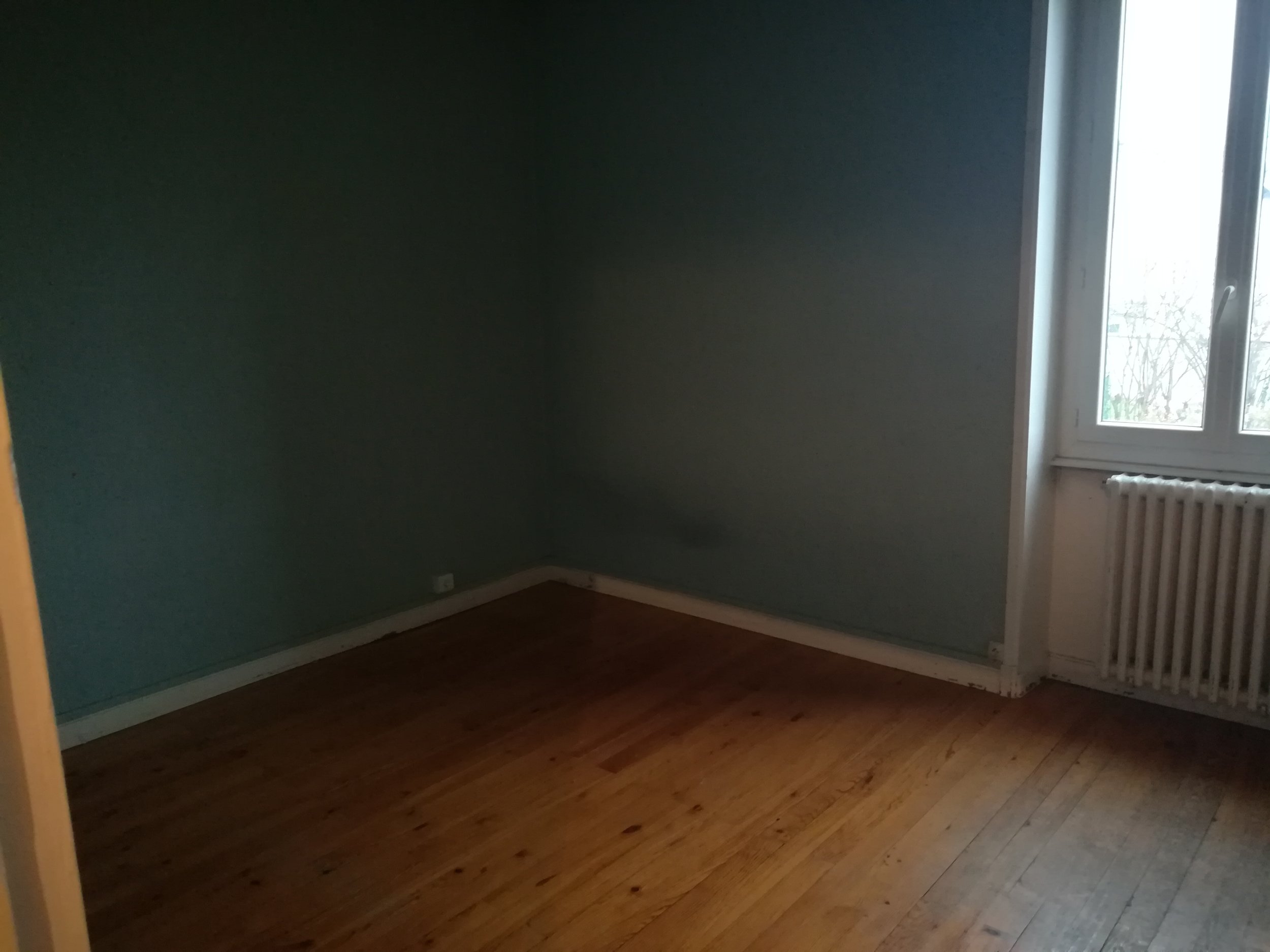 Pièce vide avec fenêtre, radiateur et parquet en bois.