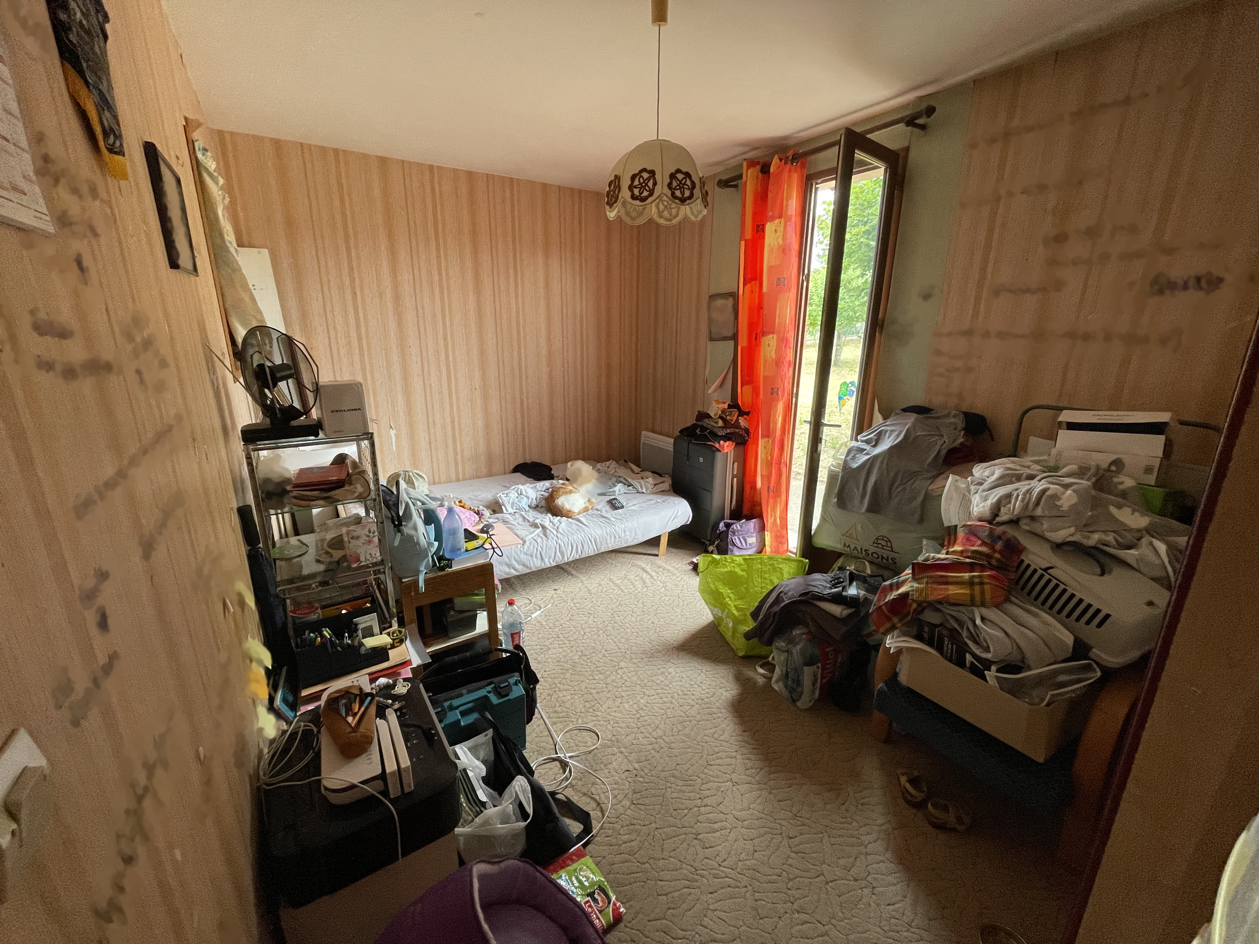 Une pièce désordonnée avec un lit couvert de vêtements, une étagère encombrée, des sacs et des objets dispersés, une porte-fenêtre laissant entrer la lumière et vue sur un extérieur vert.