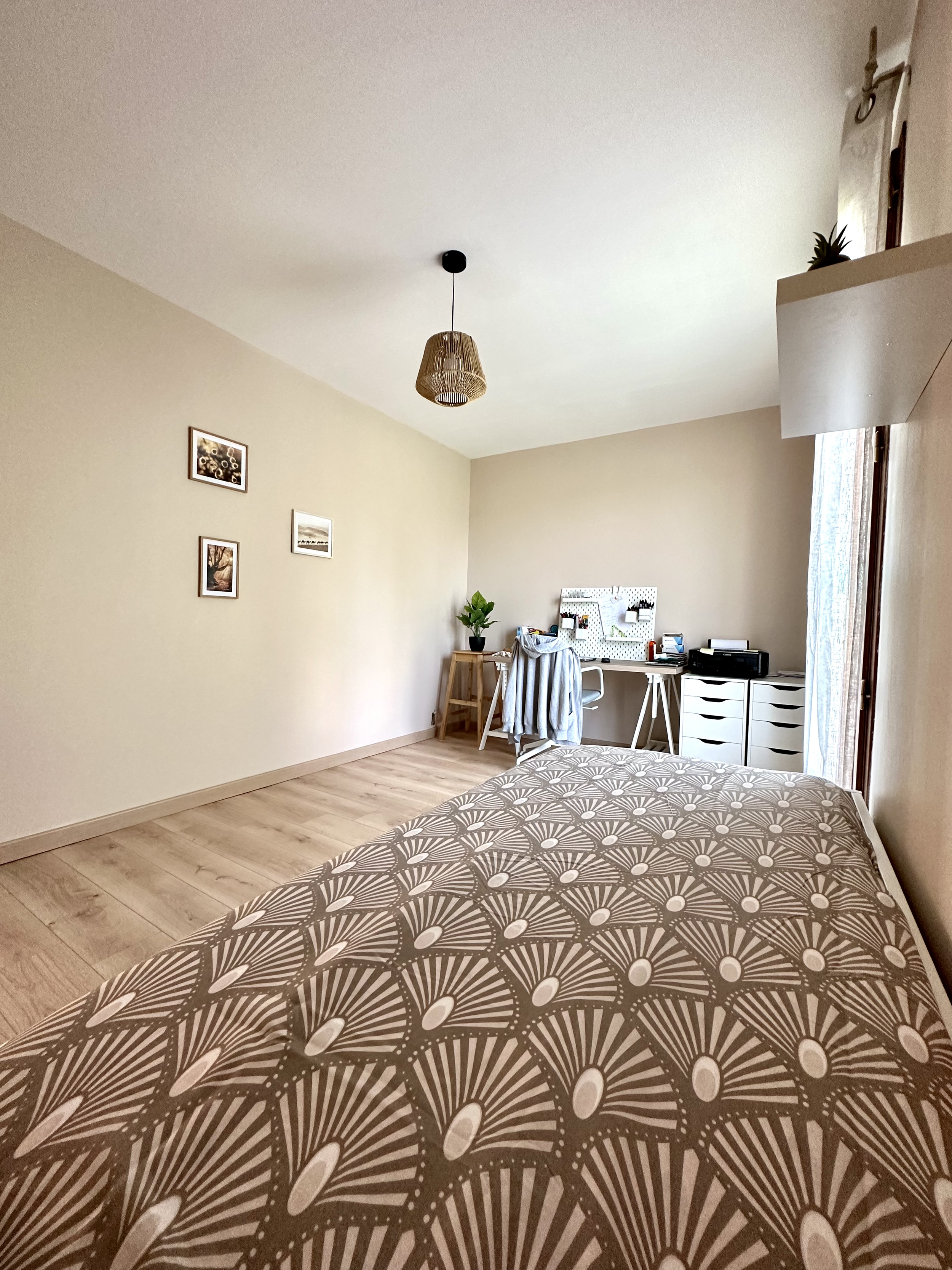 Vue partielle d'un lit avec une couette à motif géométrique beige et blanc, dans une chambre lumineuse avec un mur beige, un bureau avec des objets divers, des cadres aux murs, un plant en pot et une fenêtre avec rideau.
