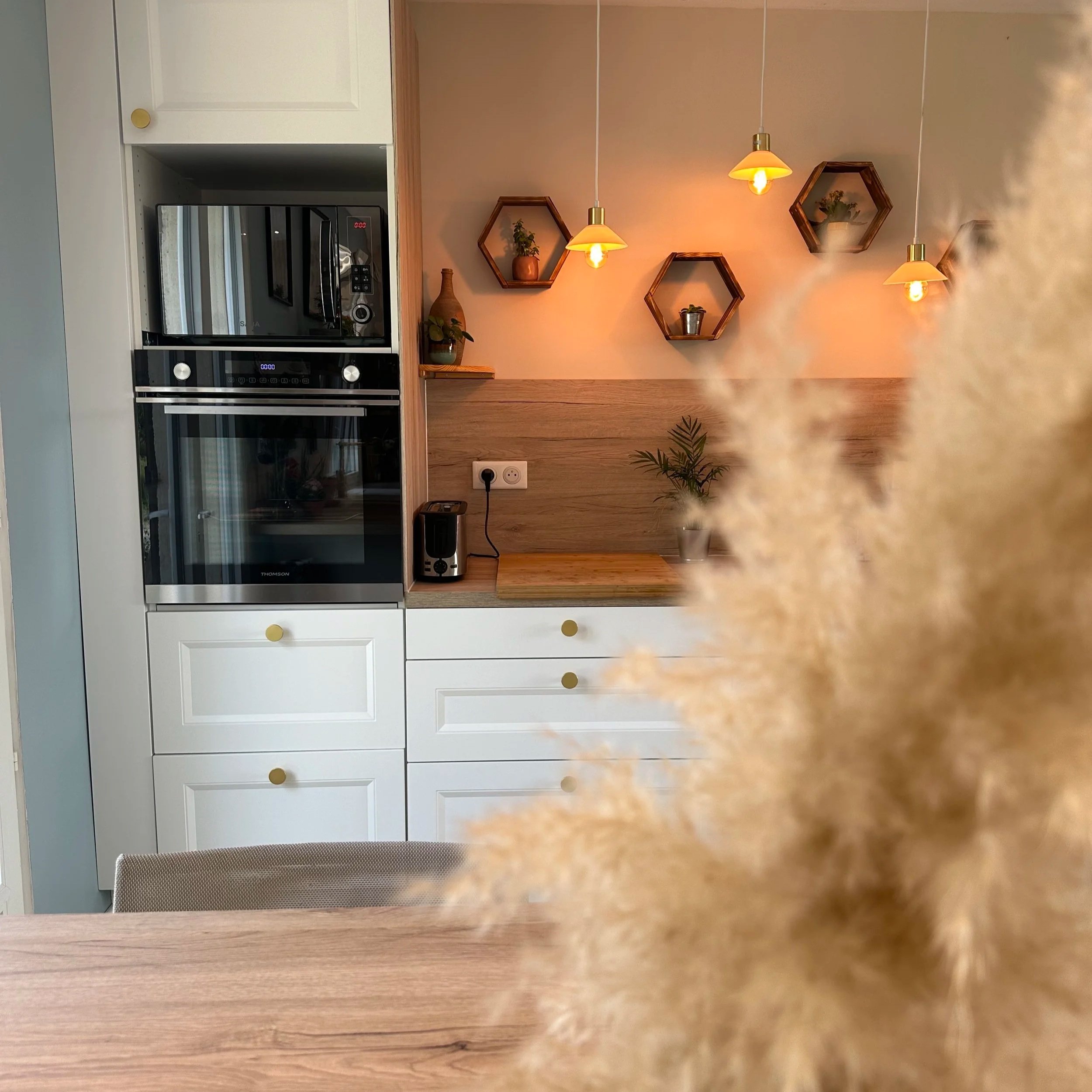 Cuisine moderne avec un mur en pierre, des étagères hexagonales en bois avec des plantes vertes, un micro-ondes noir, un four intégré, des lumières suspendues avec ampoules nues, une plante en pot, une prise électrique, un comptoir en bois et un tabouret avec un siège en tissu beige.