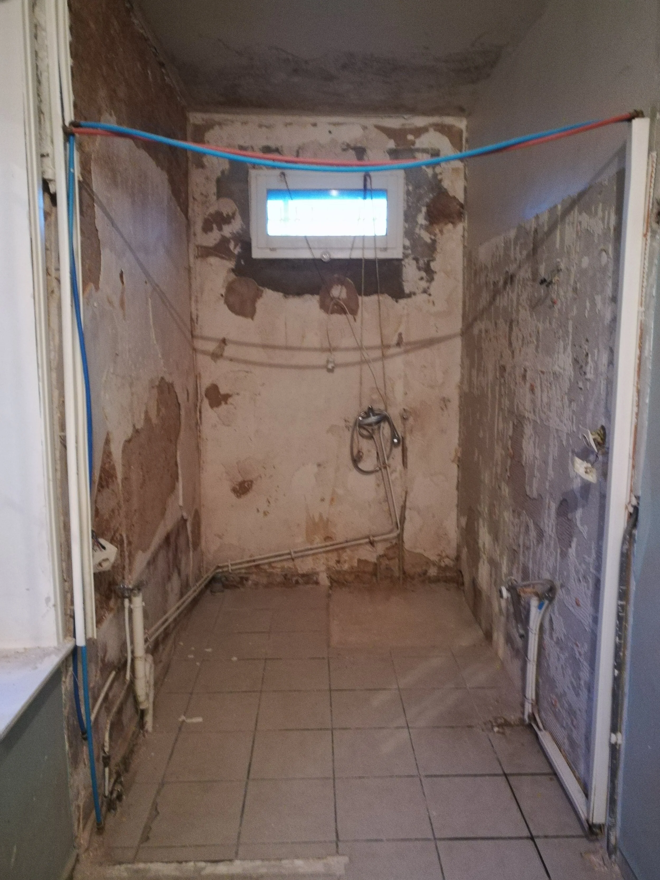 Une pièce en rénovation avec murs en enduit et câbles électriques apparents. Il y a une petite fenêtre au fond et des tuyaux au sol.