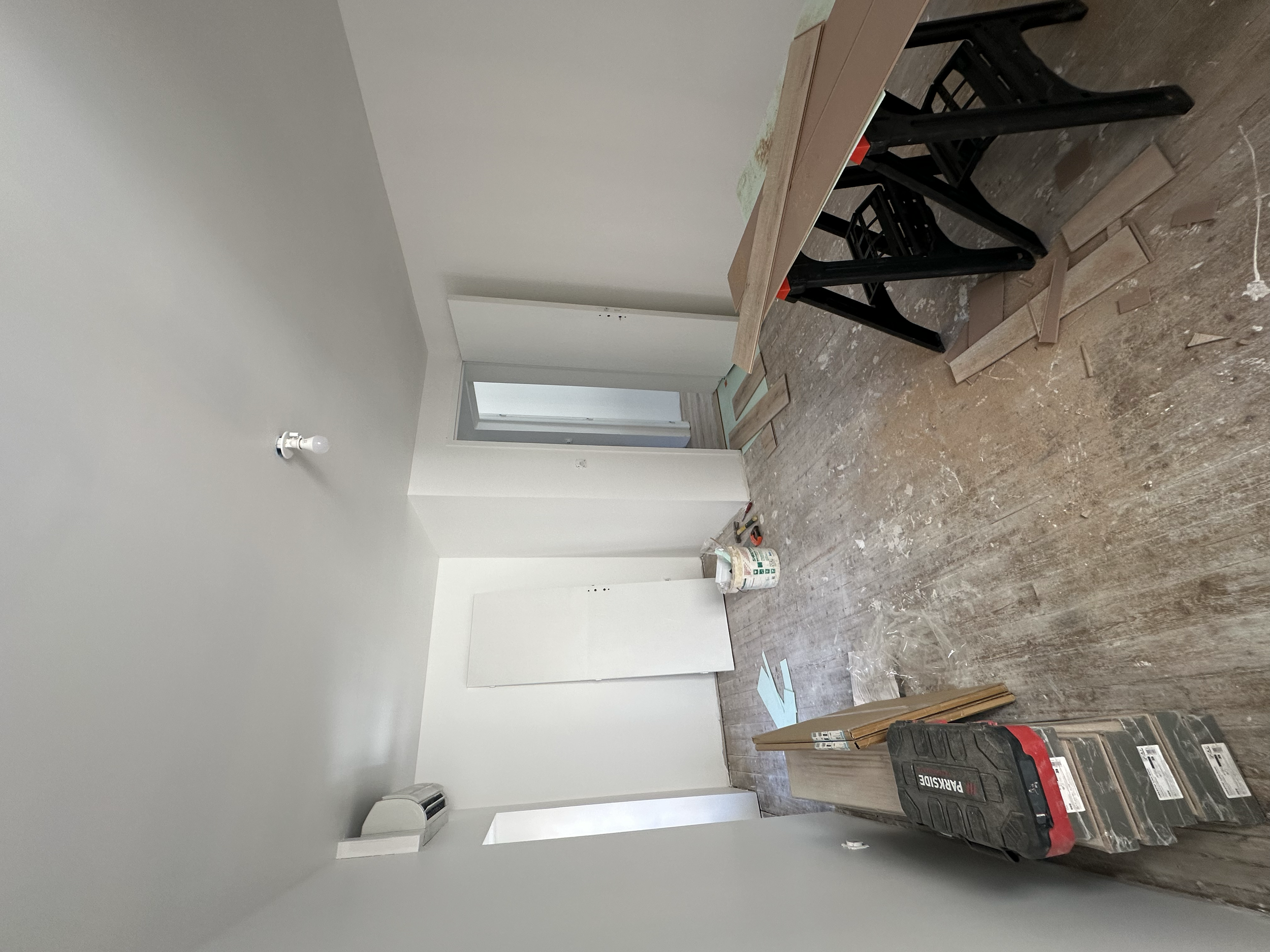 Une pièce en cours de rénovation avec des murs blancs, un sol en parquet clair, du matériel de construction, y compris un coffre à outils et des planches de bois, et des feuilles de papier sur le sol.