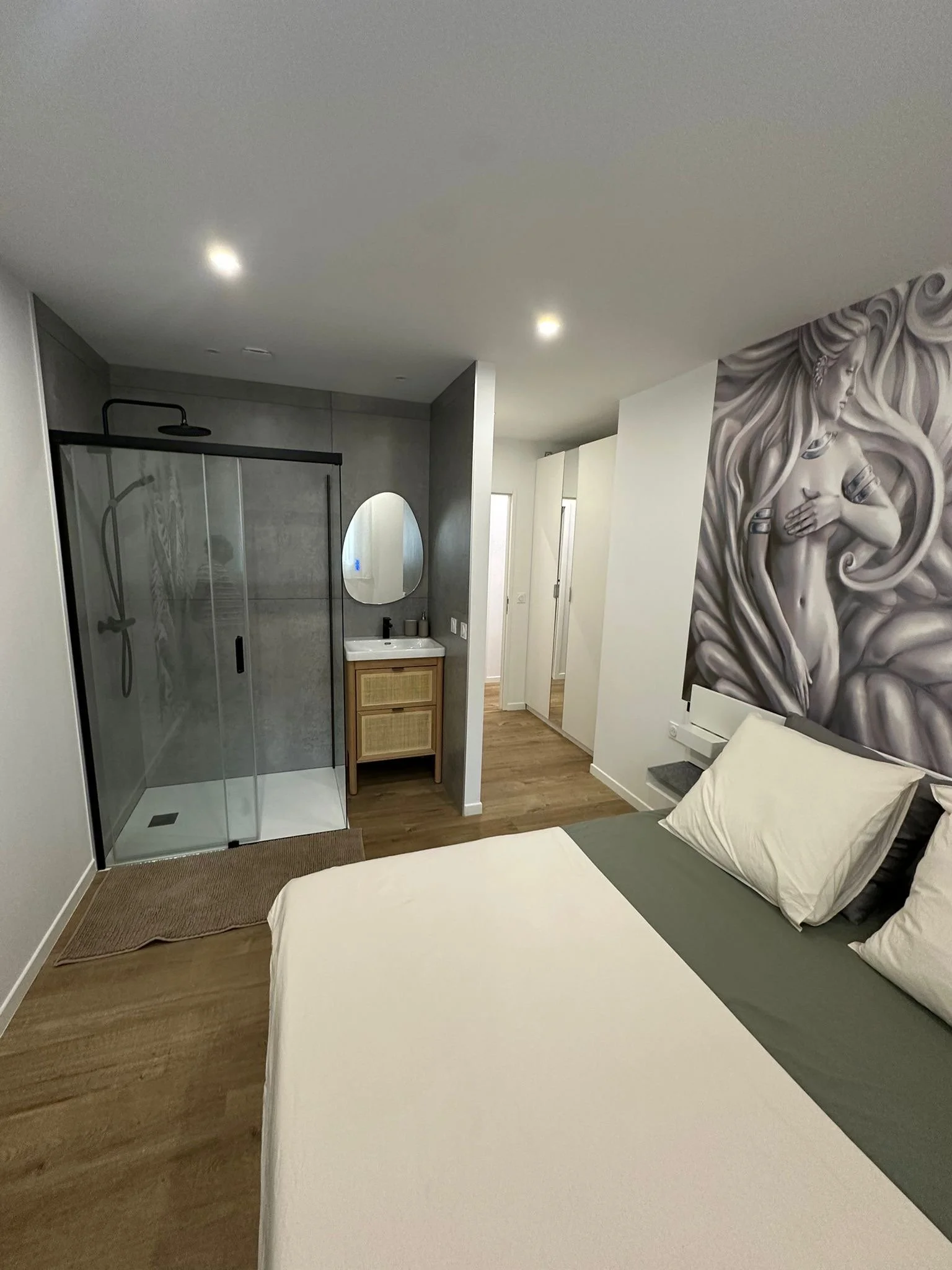 Suite parentale, chambre avec lit, murs gris, un mur avec une peinture d'une femme, salle de bain avec douche à l'italienne, miroir rond, meubles en bois, sol en bois, une petite table de nuit.