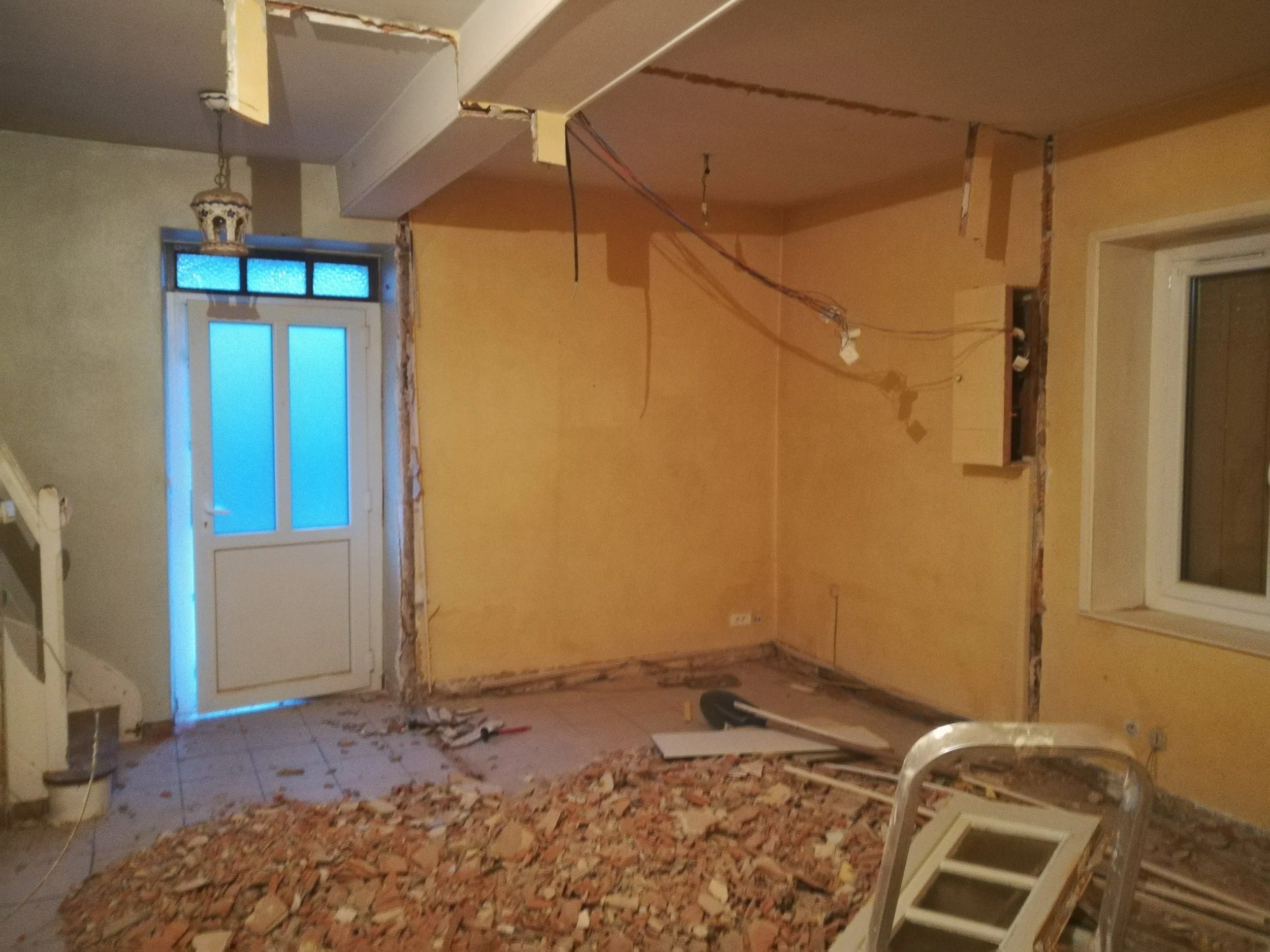 Une pièce en cours de rénovation avec un mur partiellement démoli, des débris de briques au sol, une fenêtre et un radiateur préparant à des travaux de reconstruction.