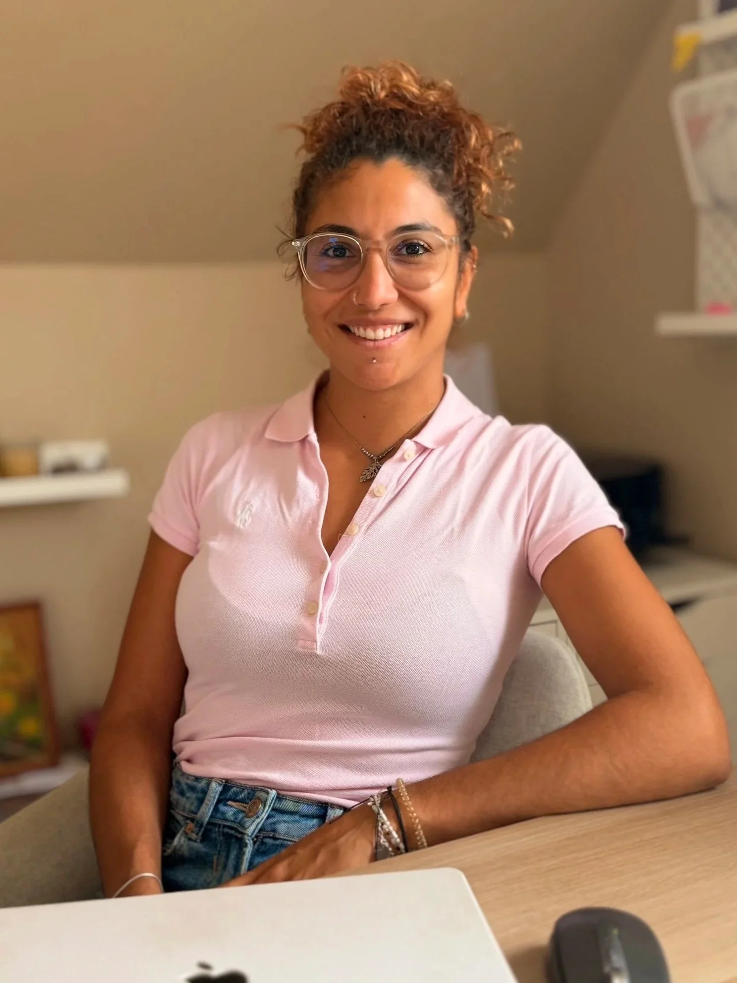 Portrait d’une femme souriante portant des lunettes et un polo rose, assise à un bureau avec un ordinateur portable.