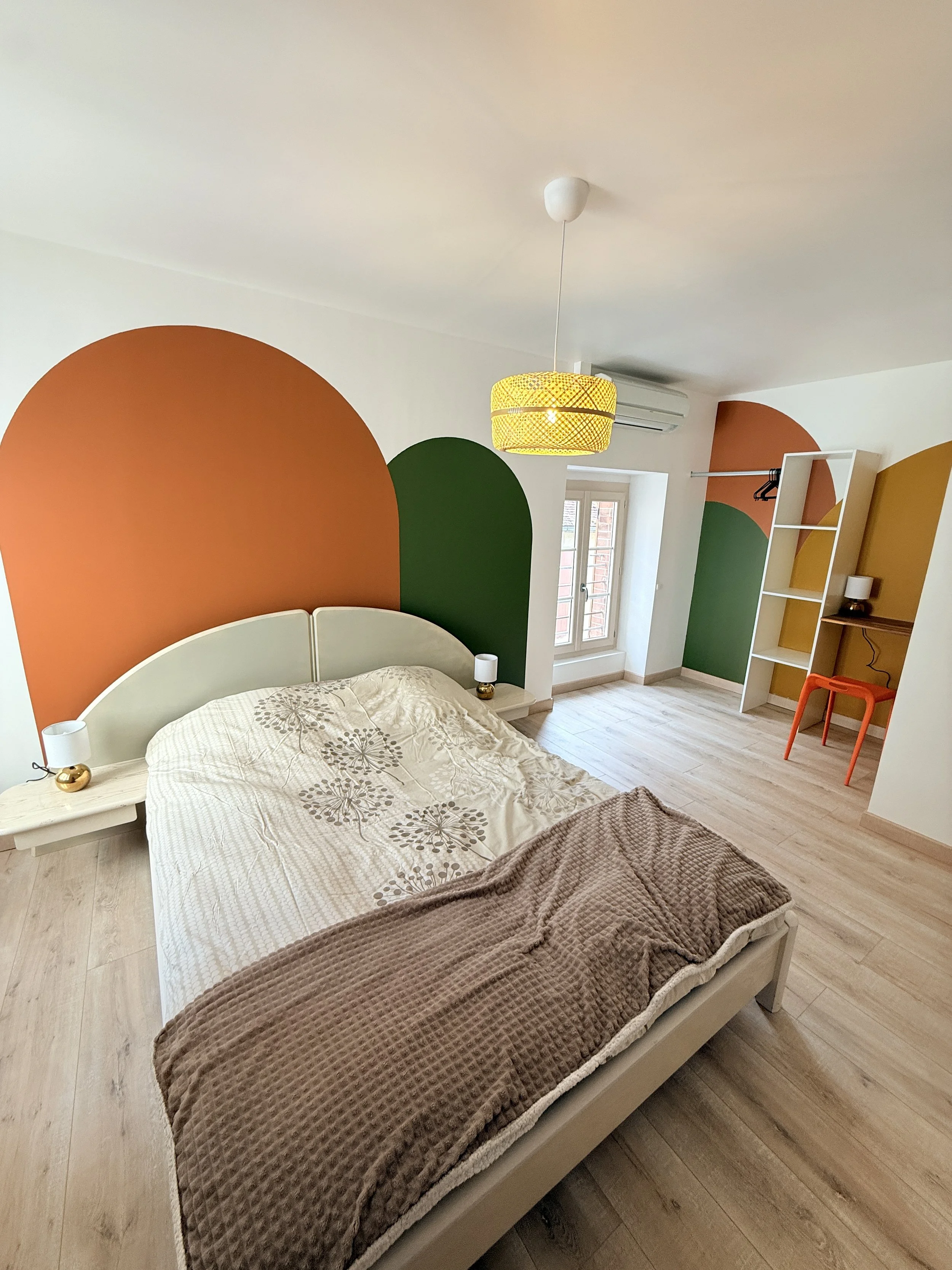Une chambre avec un lit, deux lampes de chevet, un mur avec des dessins en forme d'arcs orange, vert, jaune, une fenêtre, un climatiseur, une étagère blanche et une chaise orange.