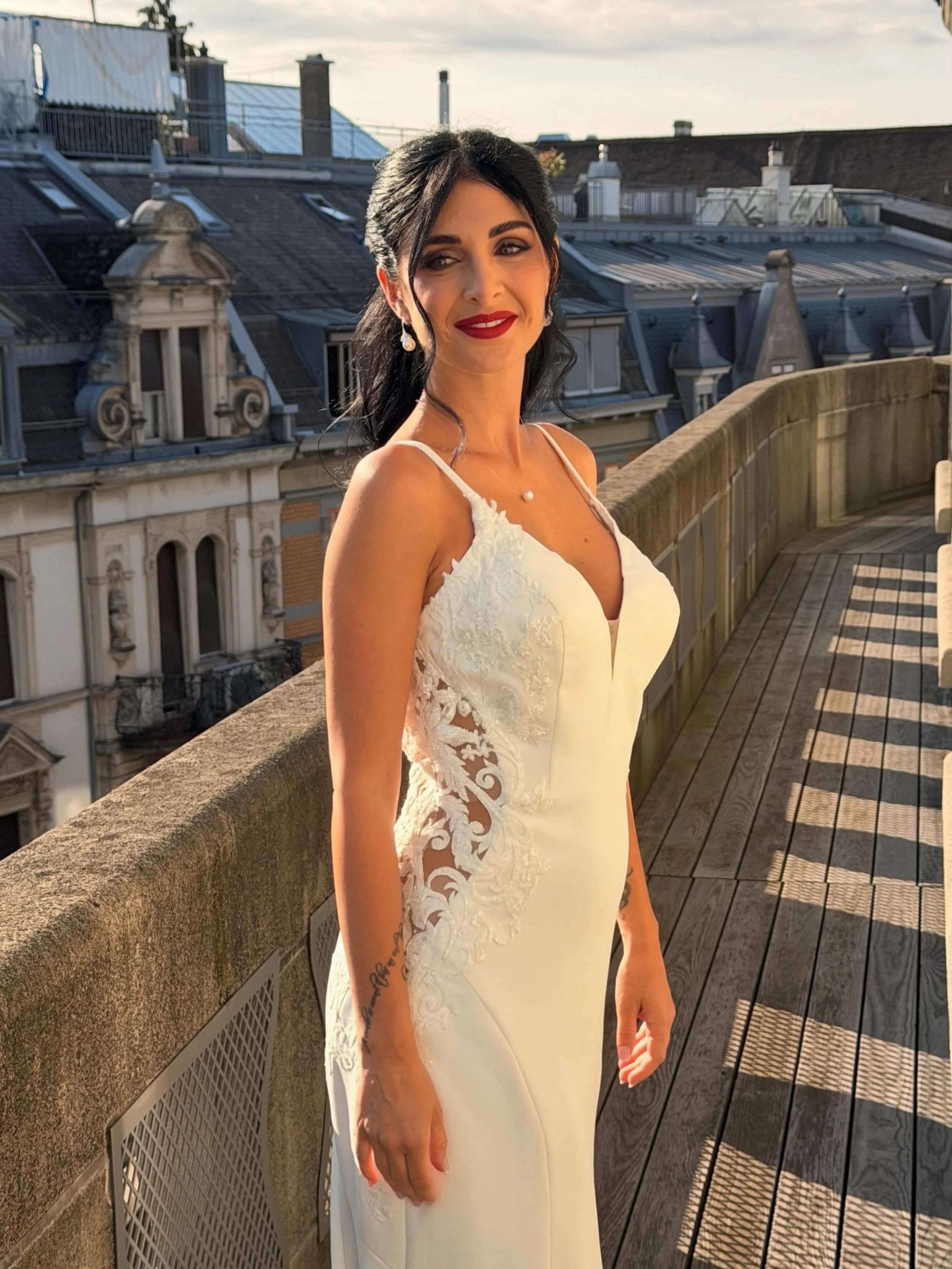 A woman in a white dress with lace details standing on a rooftop terrace with detailed building rooftops in the background, during sunset. TENZHANG MUA Switzerland