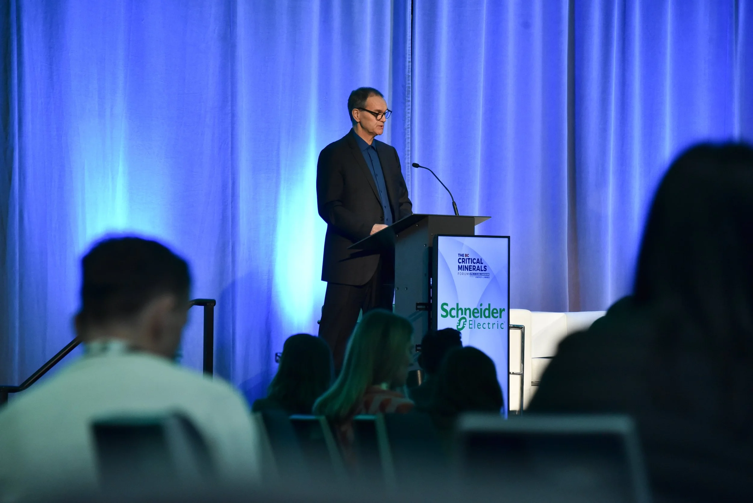 A man in a dark suit and glasses stands at a podium speaking at a conference with a large blue curtain backdrop. The display on the podium bears the logos of Schneider Electric and the BC Critical Minerals Forum.
