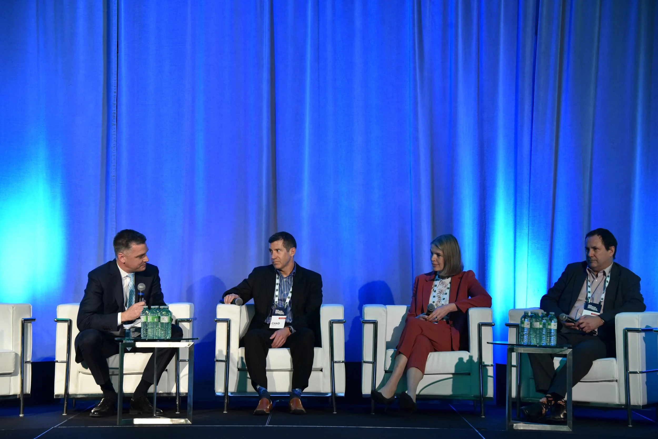 Four people sitting on stage during a panel discussion at a conference, with a blue curtain backdrop and bottled water on tables in front of them.
