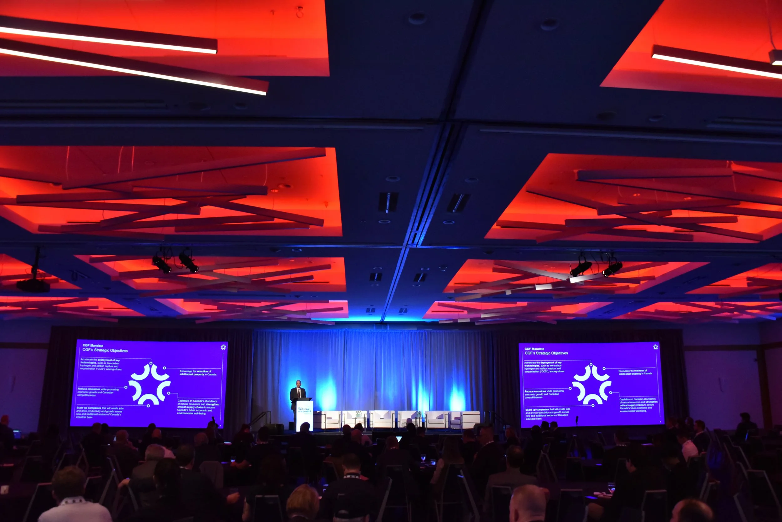 A conference or presentation event in a large ballroom with a stage, large screens with presentation slides, and an audience seated in rows. The ceiling features modern red lighting fixtures.