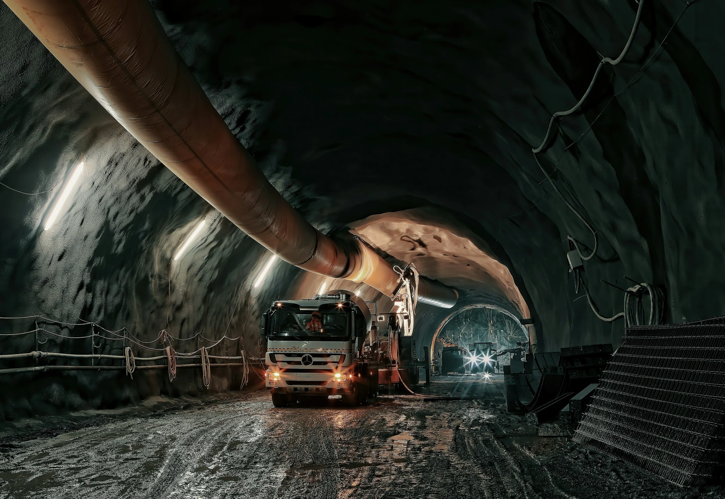 A construction vehicle working inside an underground tunnel with large pipes, lighting, and safety barriers.