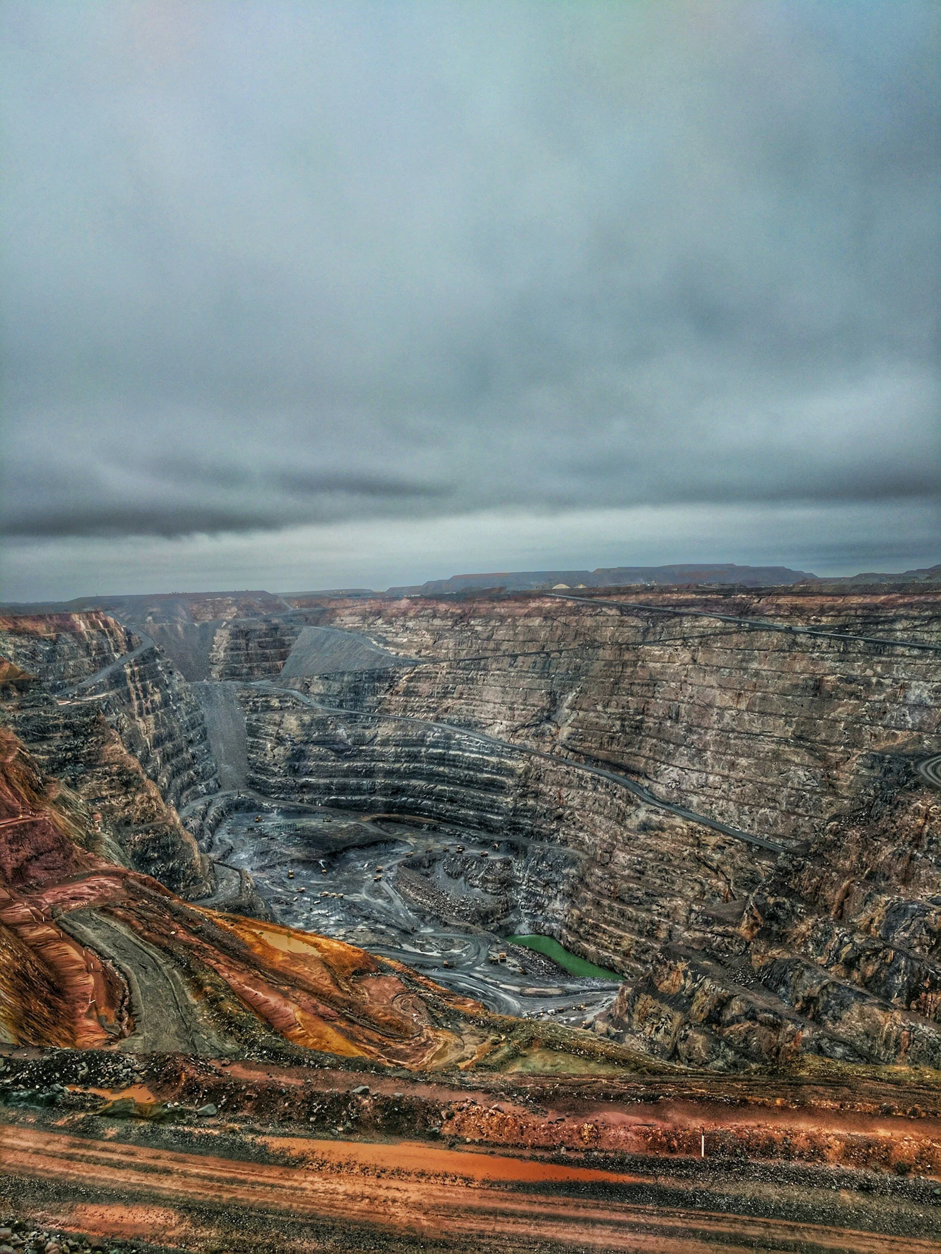 View of an expansive open-pit mine with terraced layers of earth in various shades of brown, gray, and black, and a cloudy sky overhead.