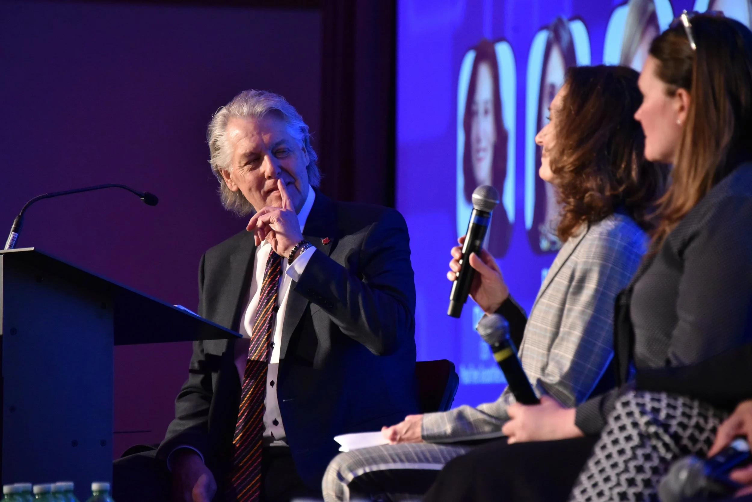 A man with gray hair in a suit and overlaying striped tie is seated at a panel, holding his finger to his lips in a gesture. A woman holding a microphone is engaging with him, and they are on a stage with a purple background and projection screens di