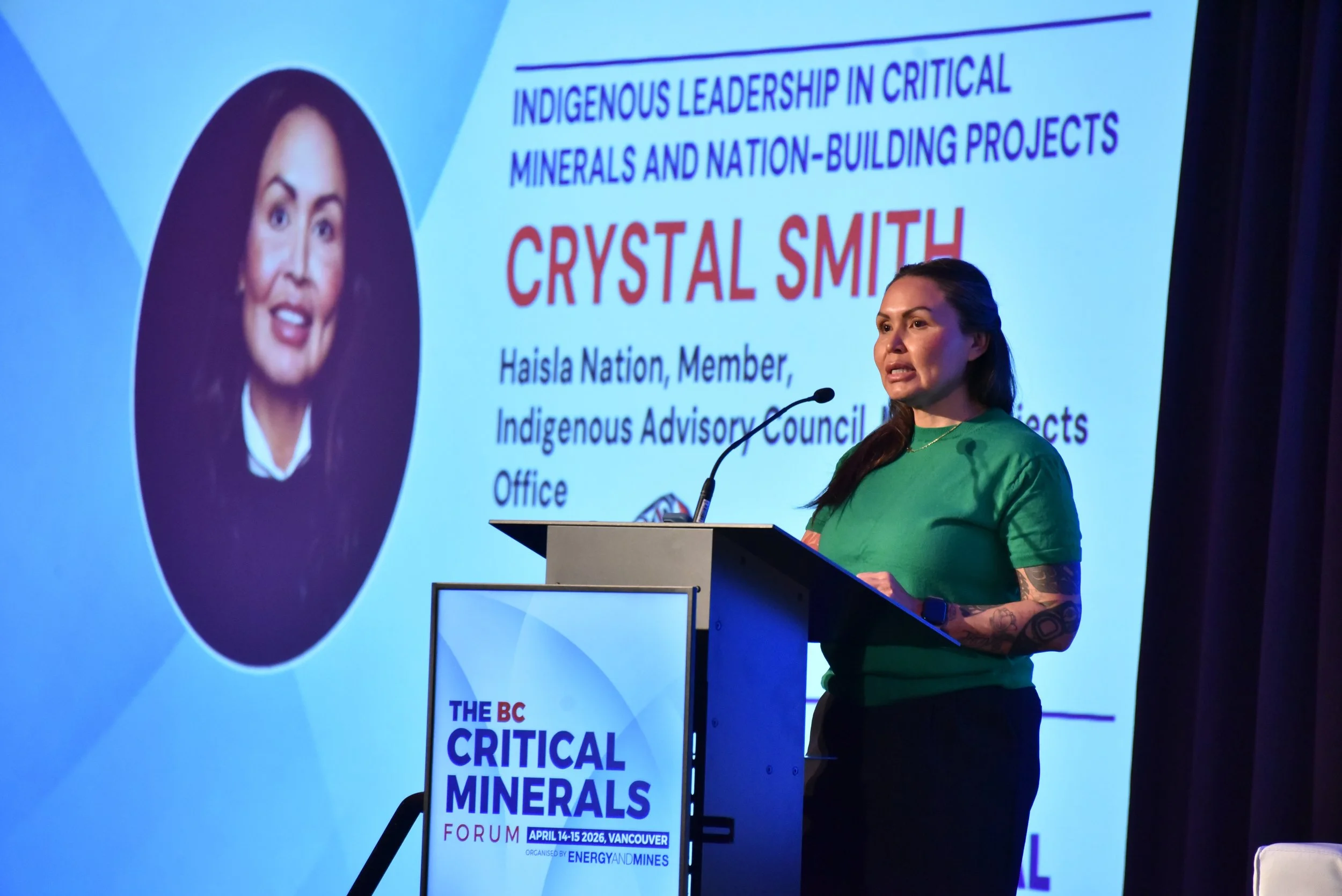 A woman with tattoos on her arm, wearing a green shirt, speaking at a podium during a conference. Behind her is a large screen displaying a presentation slide with a photo of another woman and text about indigenous leadership in critical minerals and