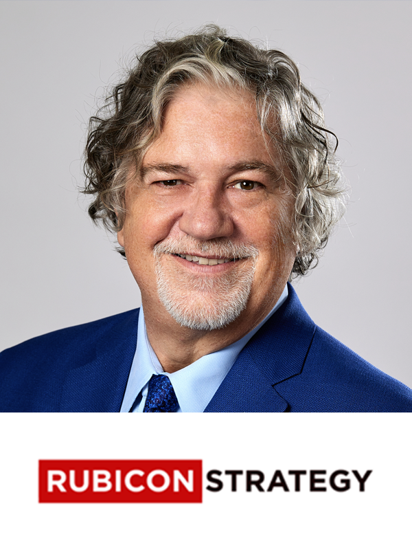 A smiling man in a blue suit and light blue shirt with a blue tie, with wavy gray hair and a beard, posing against a gray background. Below him is the logo for "Rubicon Strategy" in red and black text.