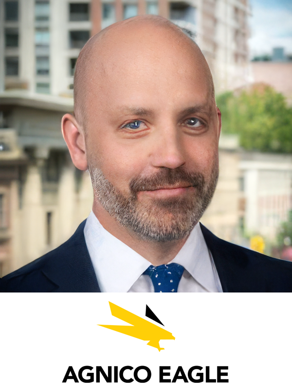 Headshot of a man with a bald head and a beard, wearing a suit and tie, with a cityscape background and the logo of Agnico Eagle at the bottom.