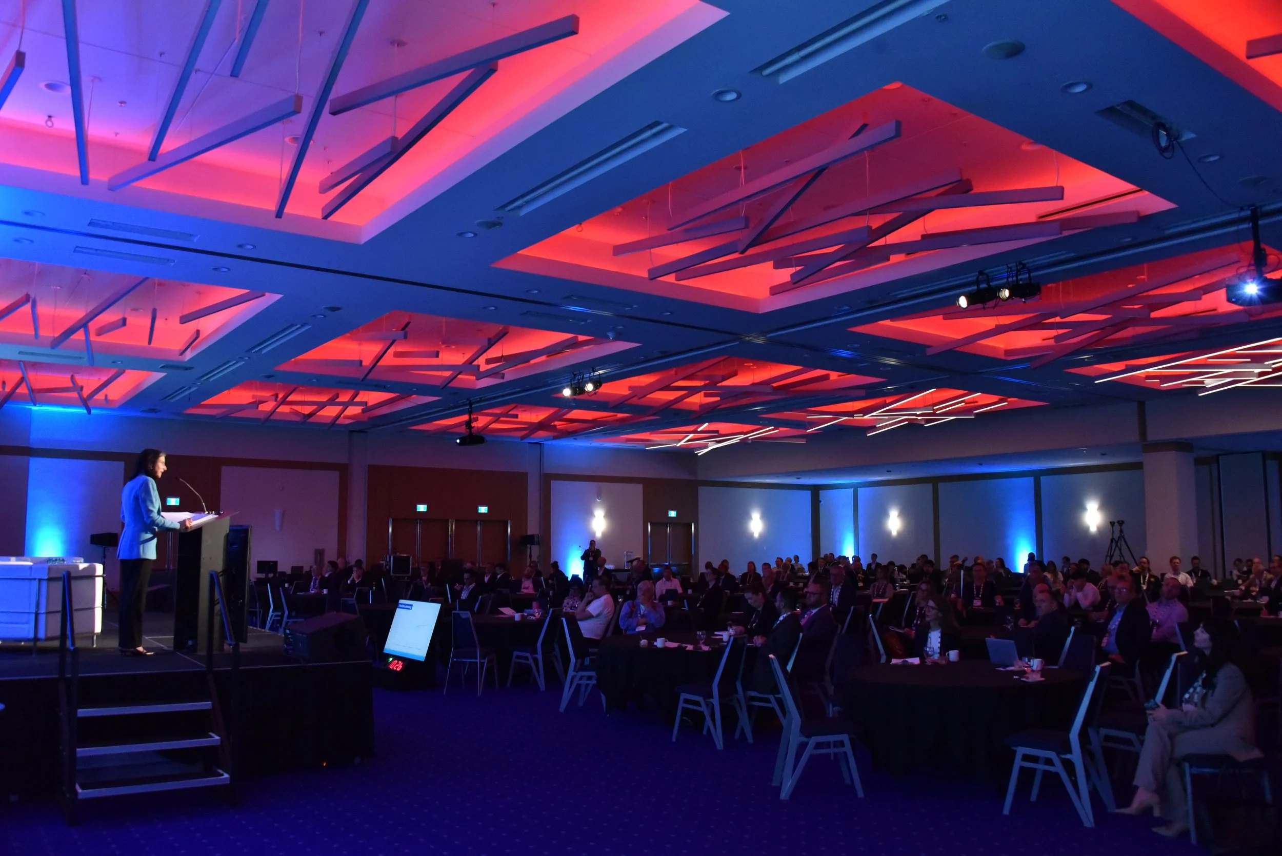 Conference room with audience seated at tables, speaker at podium on stage, illuminated ceiling with colorful lights, and projection equipment.