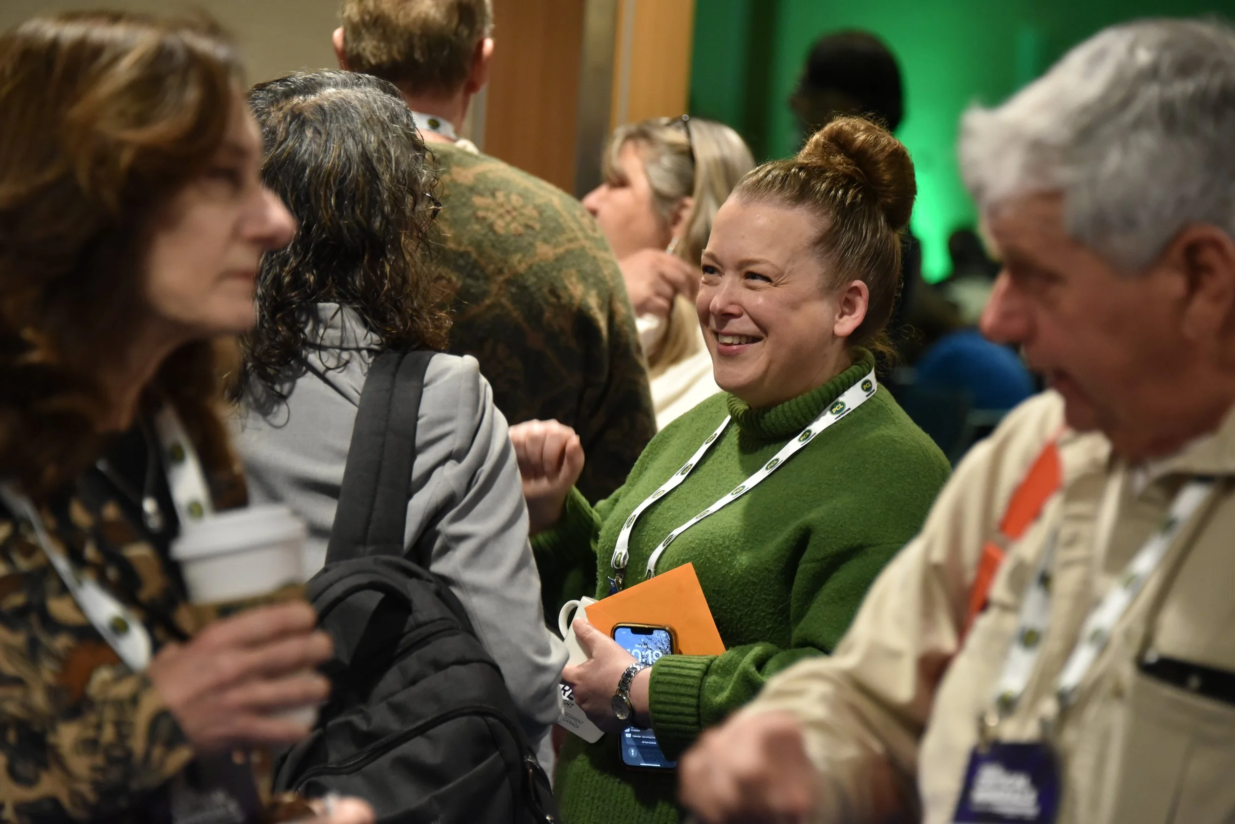 Group of people engaged in conversation at a conference or event, with a woman in green smiling and holding a phone.