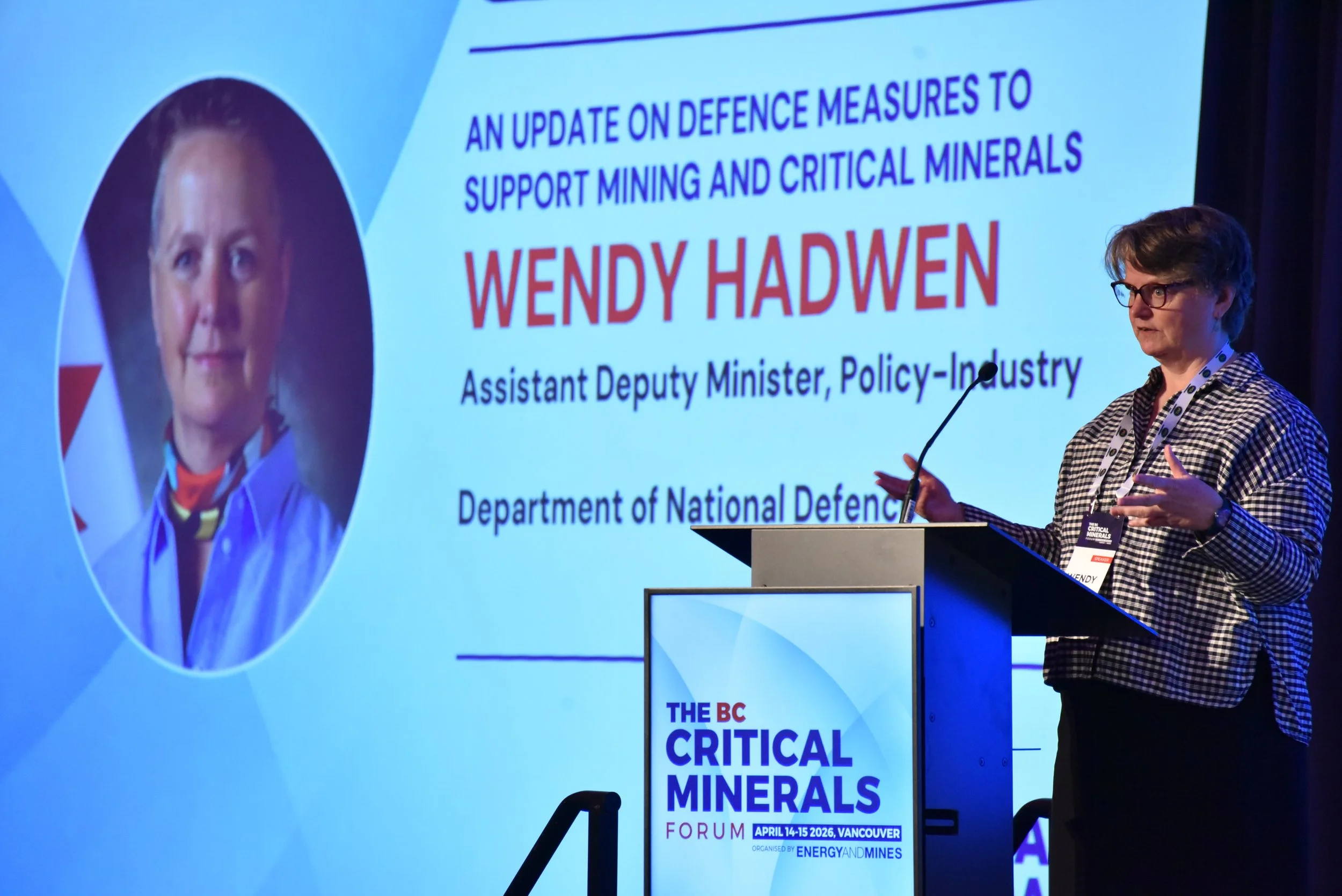A woman speaking at a conference on critical minerals, standing at a podium with a large screen behind her displaying a presentation slide. The slide features her photo, the title of the presentation about defense measures to support mining and criti