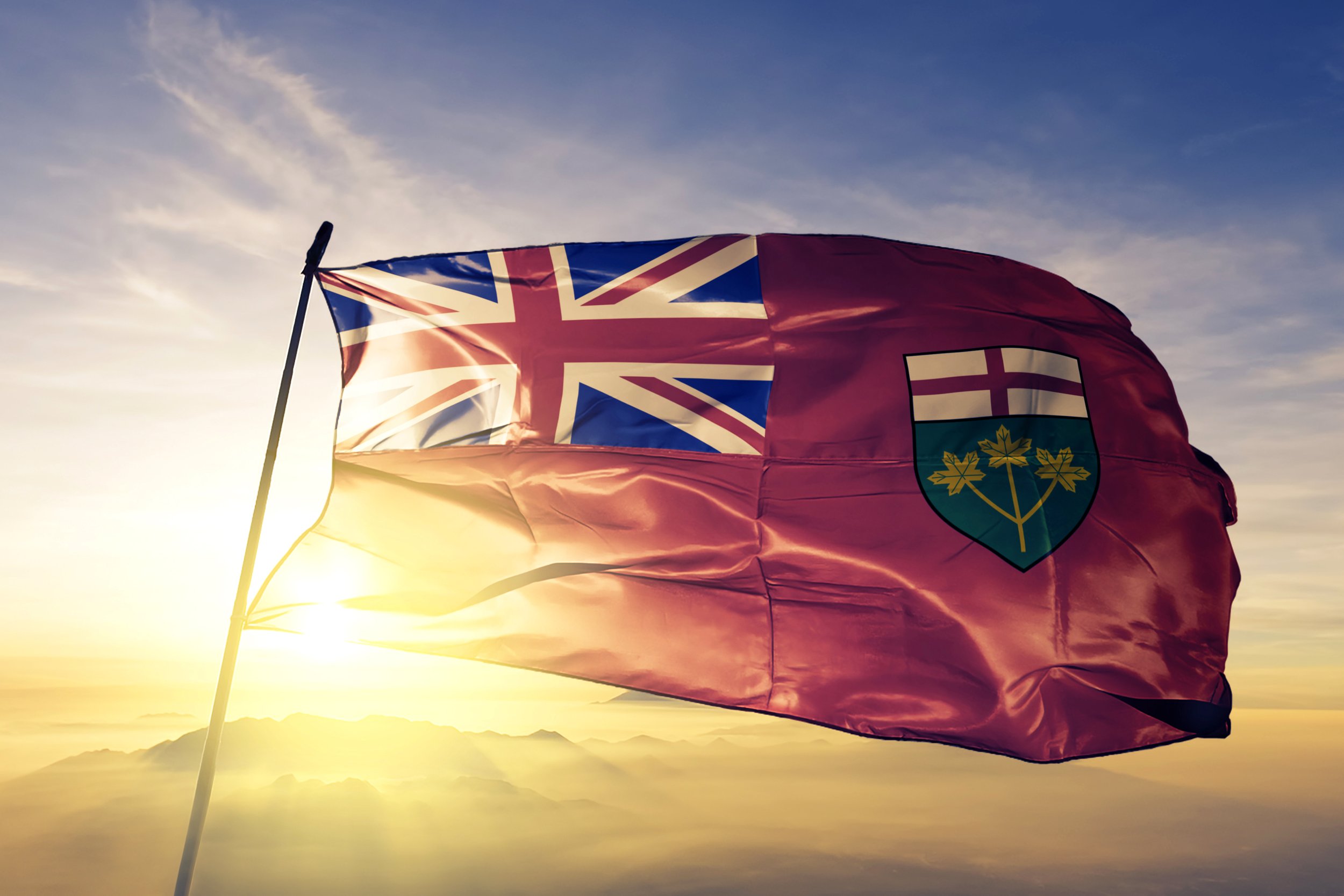 A red Ontario flag with a blue union jack corner and a shield emblem, waving in the wind against a sunset sky with clouds and mountains in the distance.