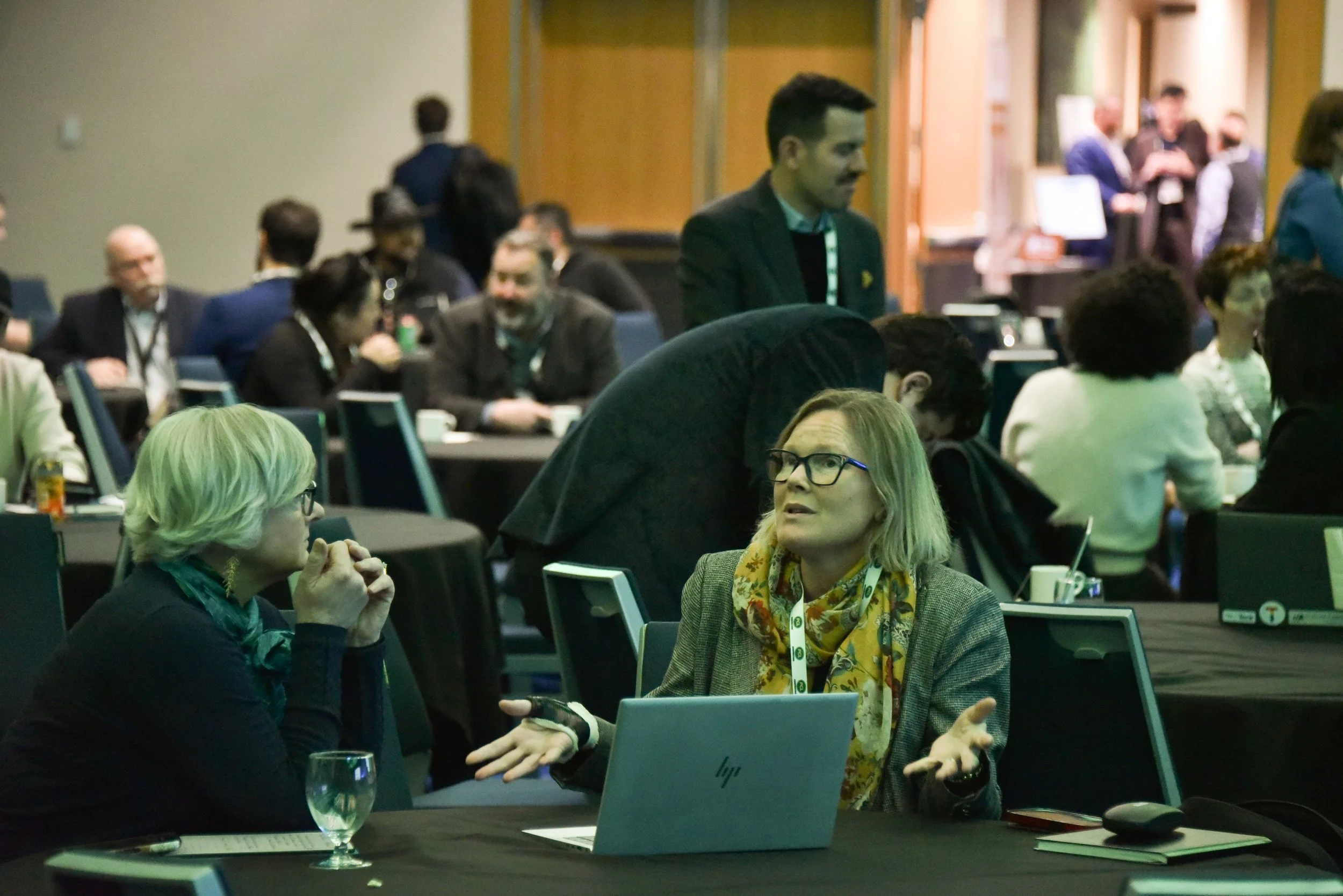 People attending a conference or business event, engaging in conversations. Some are sitting at tables with laptops and drinks, while others stand and chat in a large, well-lit room.