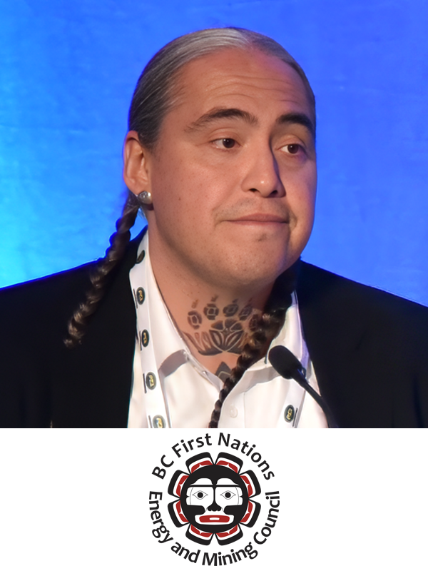 A man with long braided hair, tattoos on his neck, wearing a white shirt, black blazer, and a lanyard, speaking at a conference with a blue background, with a BC First Nations Energy and Mining Council logo at the bottom.