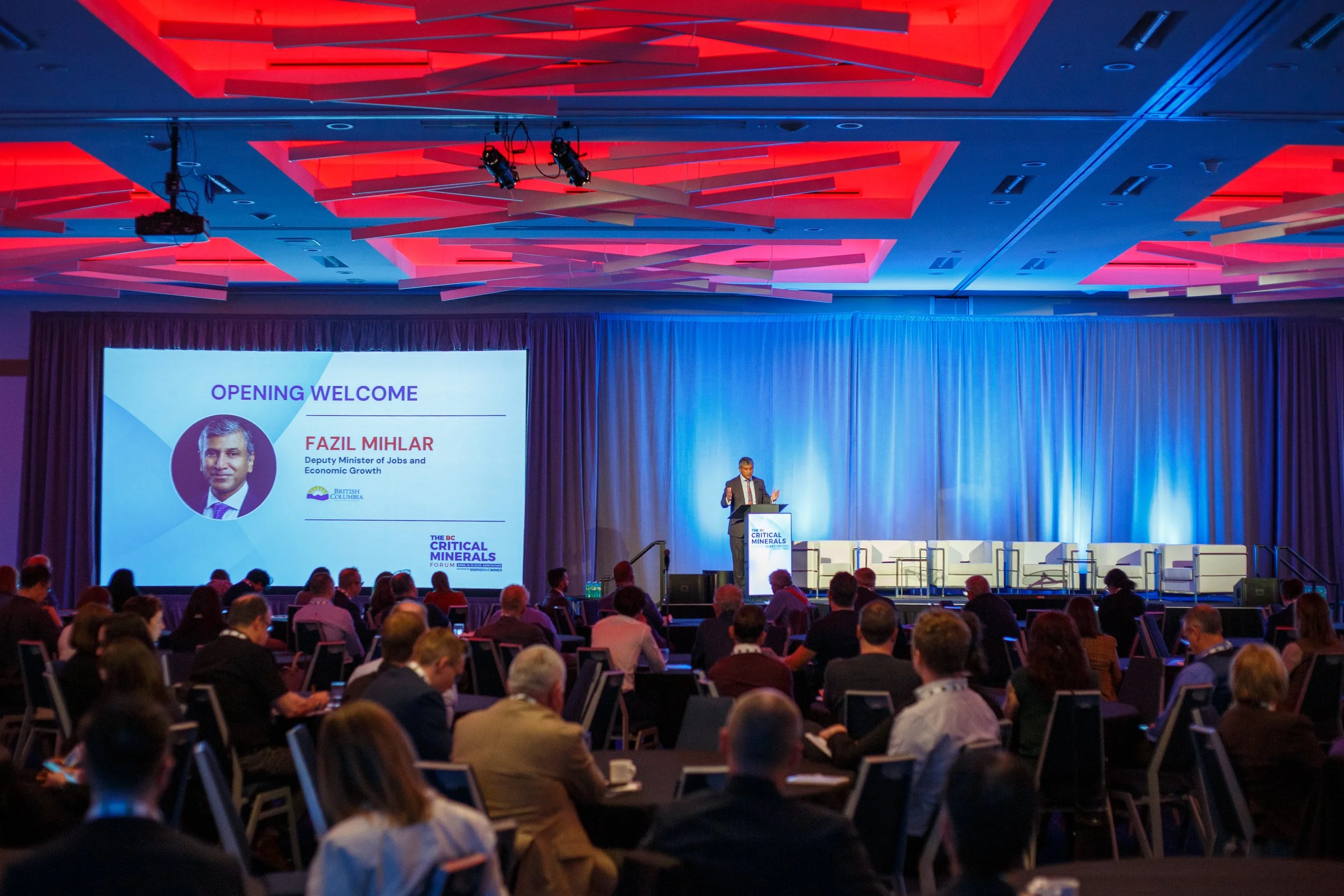 A conference room filled with attendees listening to a speaker. The stage features a large screen showing a presentation slide with the title "Opening Welcome" and a photo of Fazil Milhar, Deputy Minister of Jobs and Economic Growth. The ceiling has 