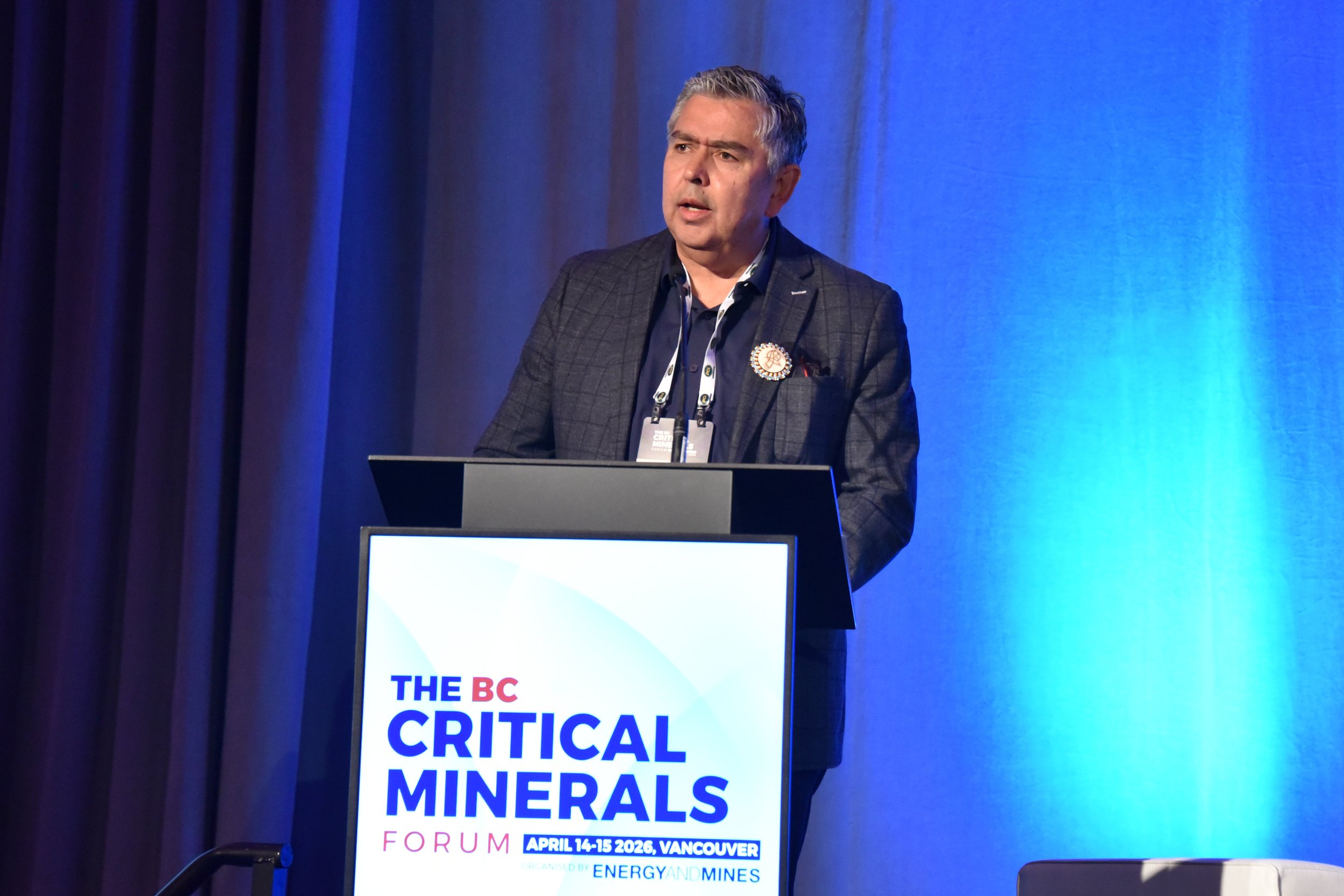 A man with gray hair standing behind a podium at a conference on critical minerals. He is wearing a dark plaid blazer and a lanyard with badges, with a serious expression. The background features blue curtains and a screen displaying the event detail