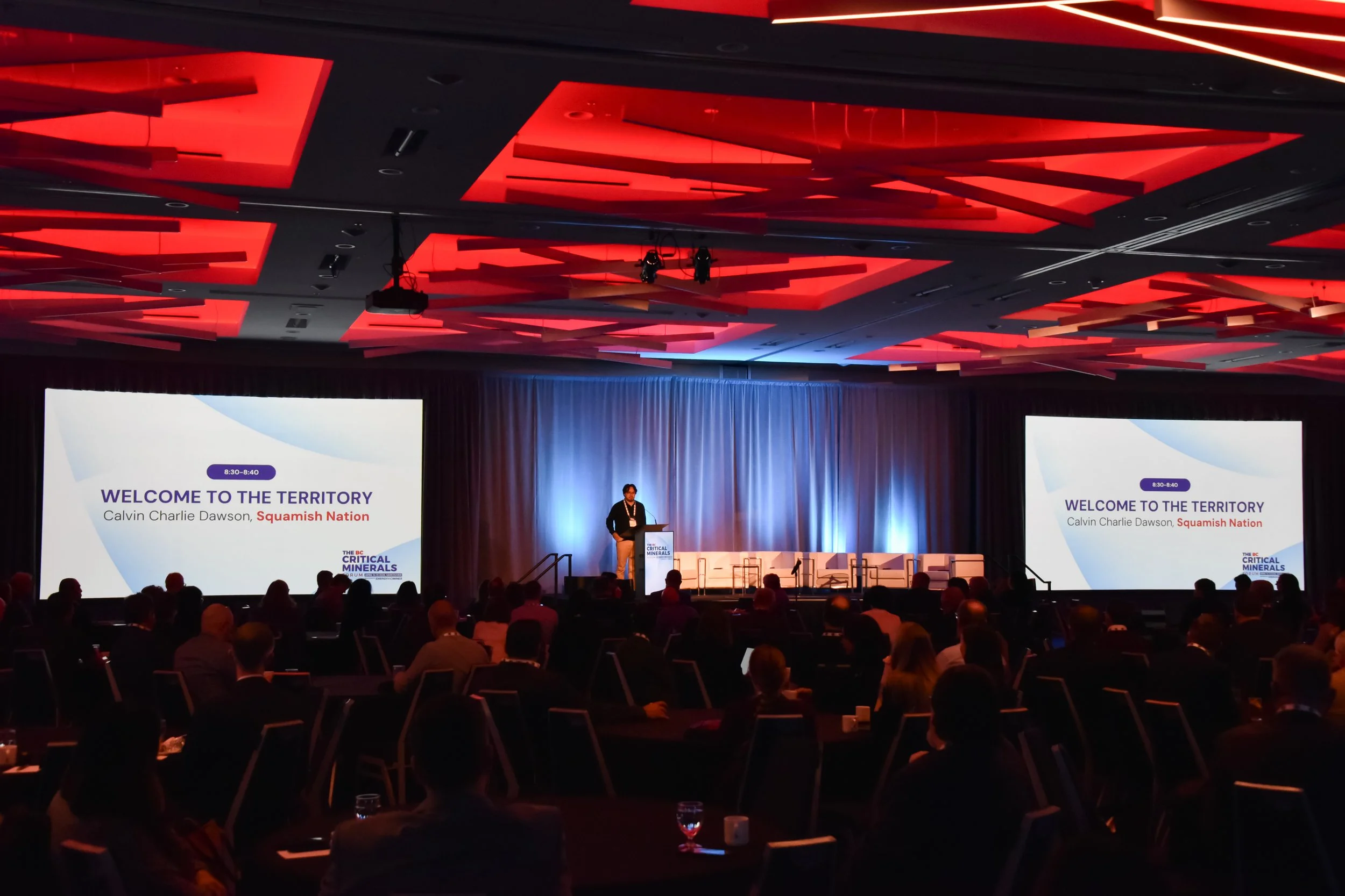 A conference room with a stage and large screens displaying the presentation slide titled 'Welcome to the Territory' by Calvin Charlie Dawson, Squamish Nation. The ceiling has red geometric lighting designs, and there are many attendees seated facing