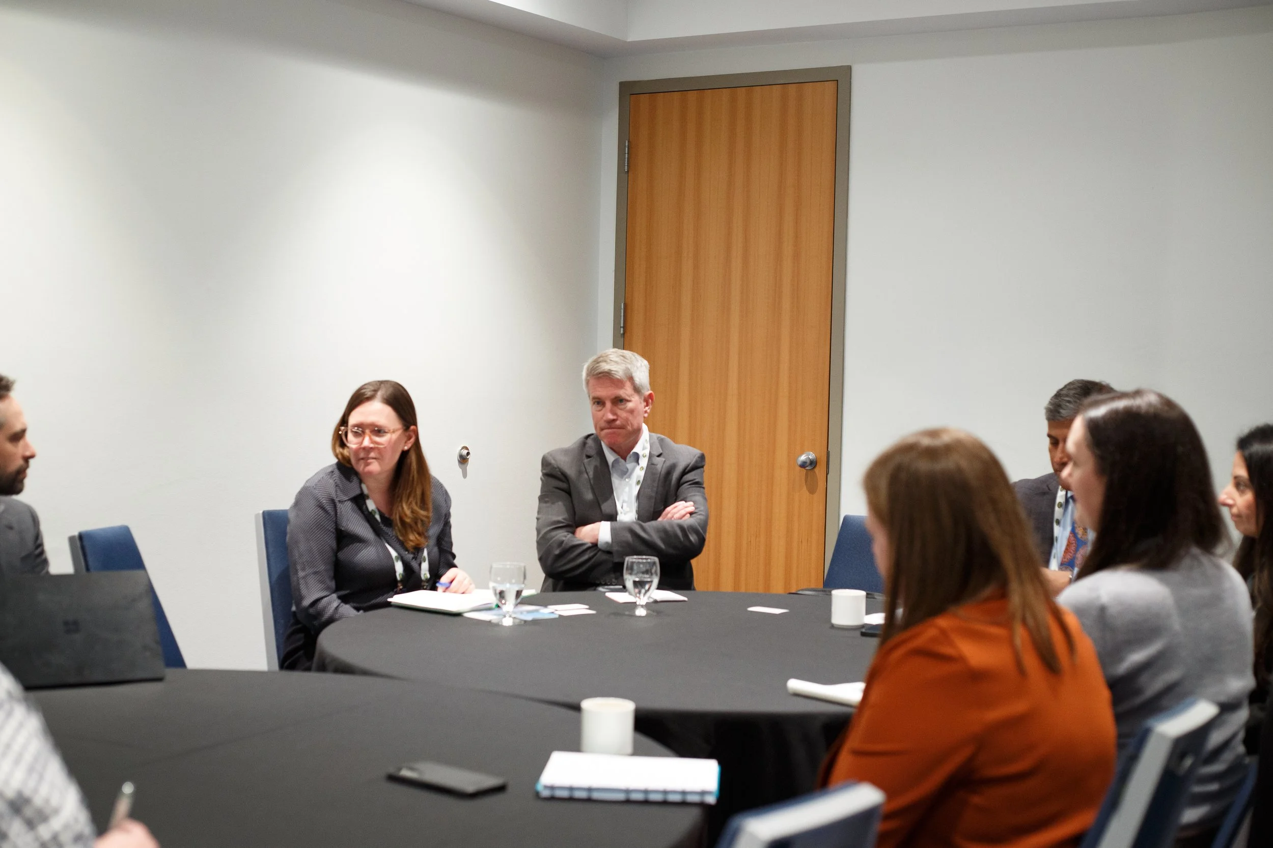 A group of diverse people sitting around a conference table, engaged in a discussion, with some taking notes and others listening attentively, in a modern conference room with a wooden door and white walls.