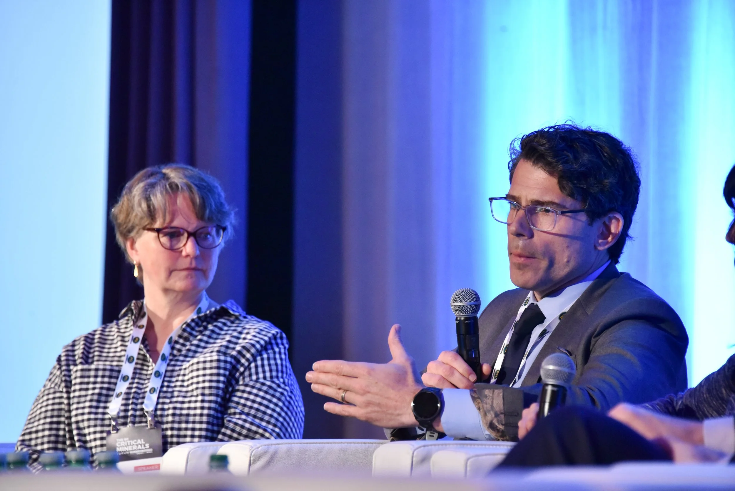 A woman and a man sitting at a panel discussion. The woman has short, light-colored hair, glasses, and is wearing a checkered shirt. The man has dark, curly hair, glasses, and a dark suit, and is holding a microphone. The background features dark cur