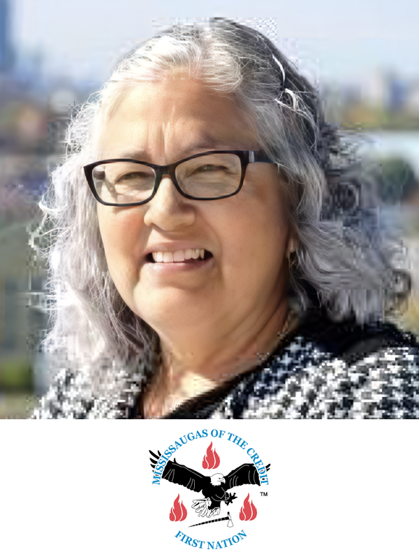 A middle-aged woman with gray hair and glasses, outdoors with a blurry cityscape background, wearing a patterned top and smiling slightly. Below her image is the logo of Mississaugas of the Credit First Nation, which features black hands, red flames, and blue text.