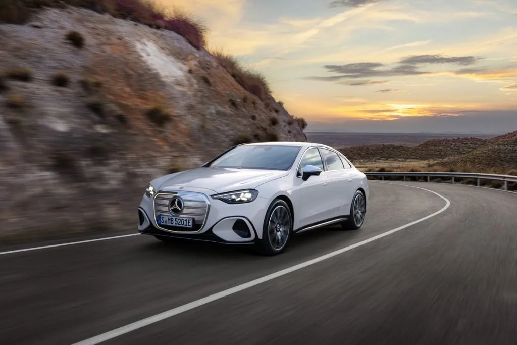 A silver Mercedes-Benz sedan driving on a winding mountain road at sunset with hills and a cloudy sky in the background.