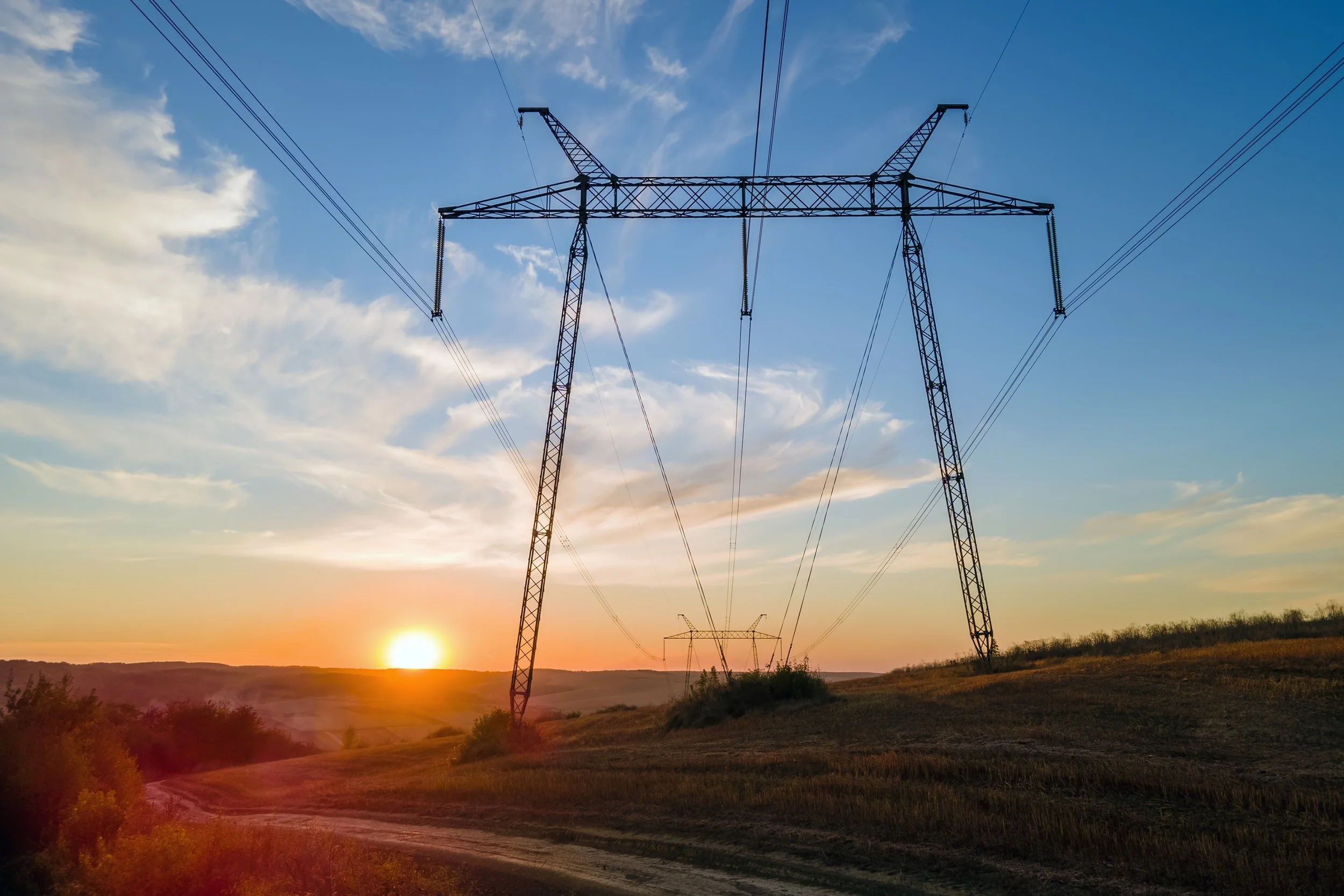 Sunset over a rural landscape with power lines and transmission towers stretching into the distance.