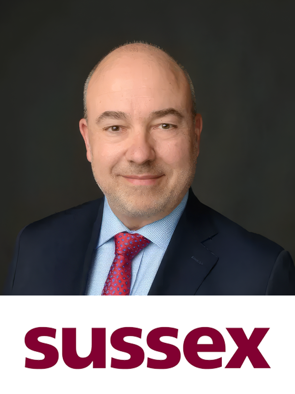 A professional headshot of a smiling middle-aged man with a bald head, wearing a navy blazer, light blue shirt, and red patterned tie, against a dark background. The word "Sussex" is written in bold maroon letters below the image.
