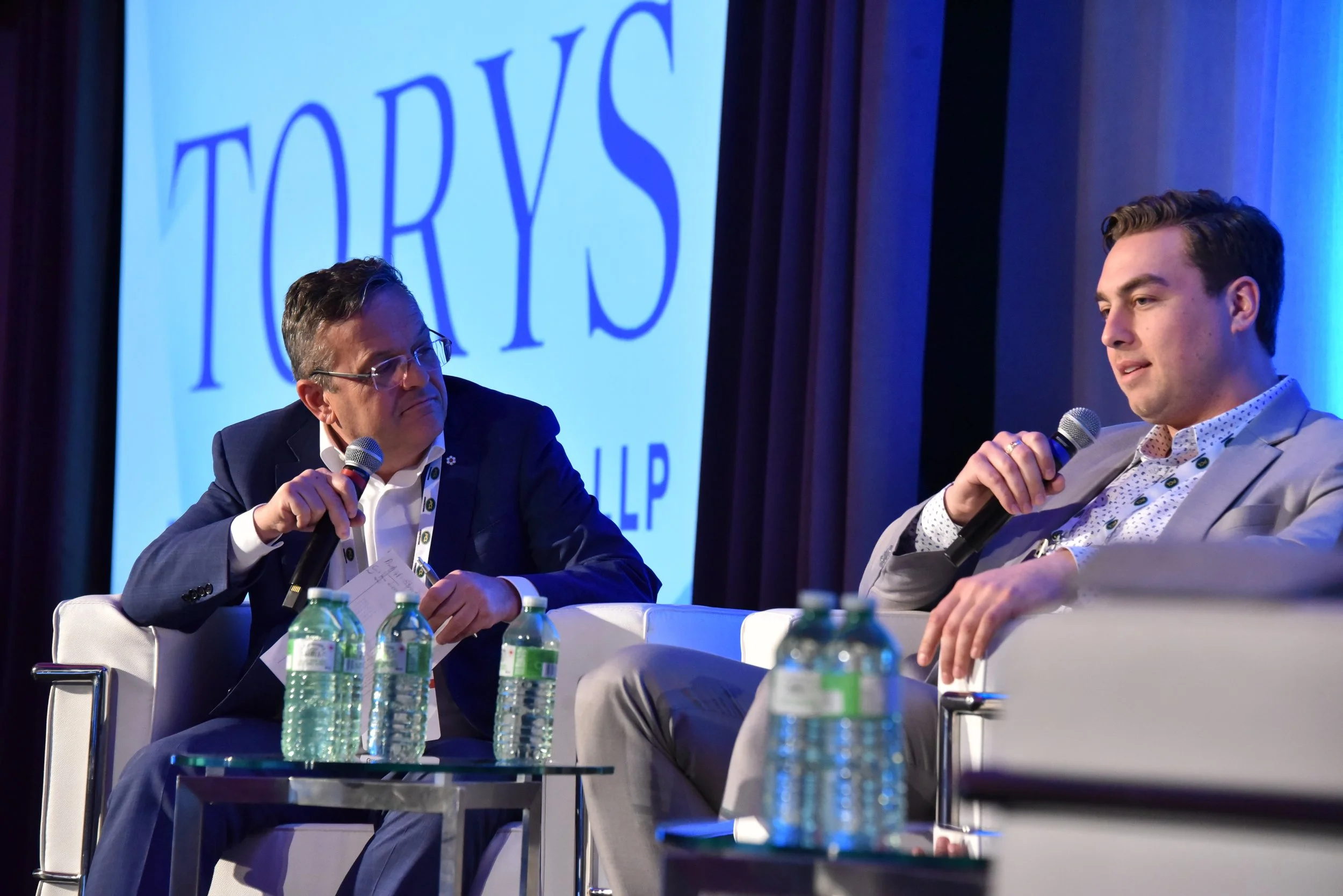 Two men sitting on stage and speaking into microphones during a panel discussion, with a large blue 'TOYS' sign in the background, and several water bottles on the table in front of them.
