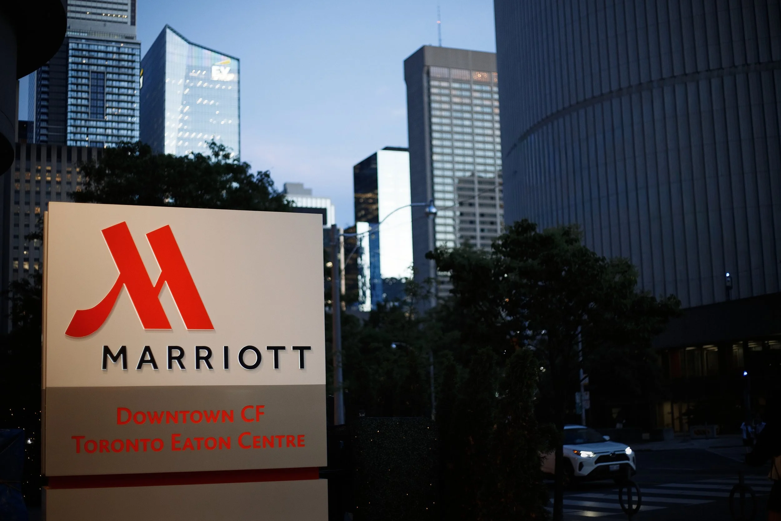 A city street scene at dusk with tall glass buildings in the background and a large sign for Marriott hotel in the foreground. The sign indicates downtown Toronto and the Eaton Centre.